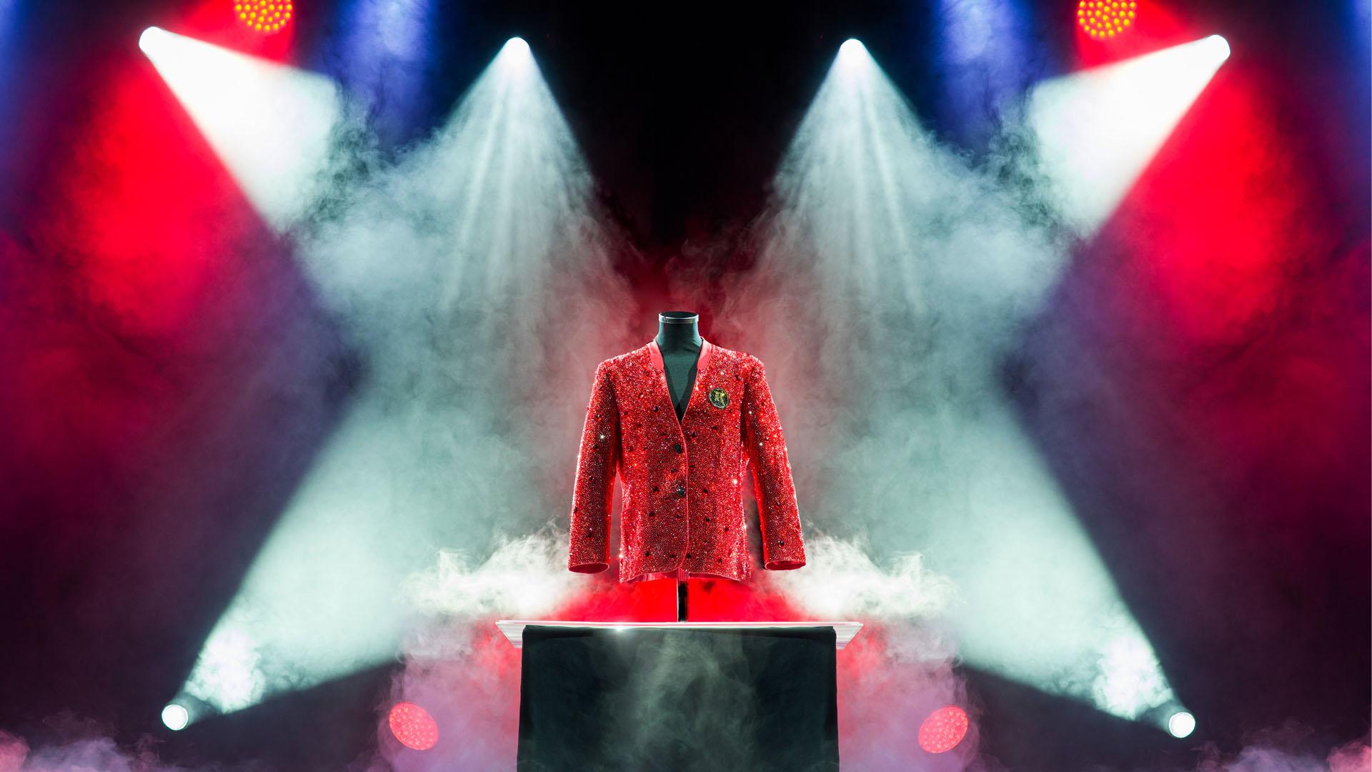 A red crystal-covered blazer on a black mannequin displayed on an illuminated pedestal, bathed in dramatic stage lighting with red, blue, and white spotlights converging from above. Thick atmospheric fog surrounds the base of the display.