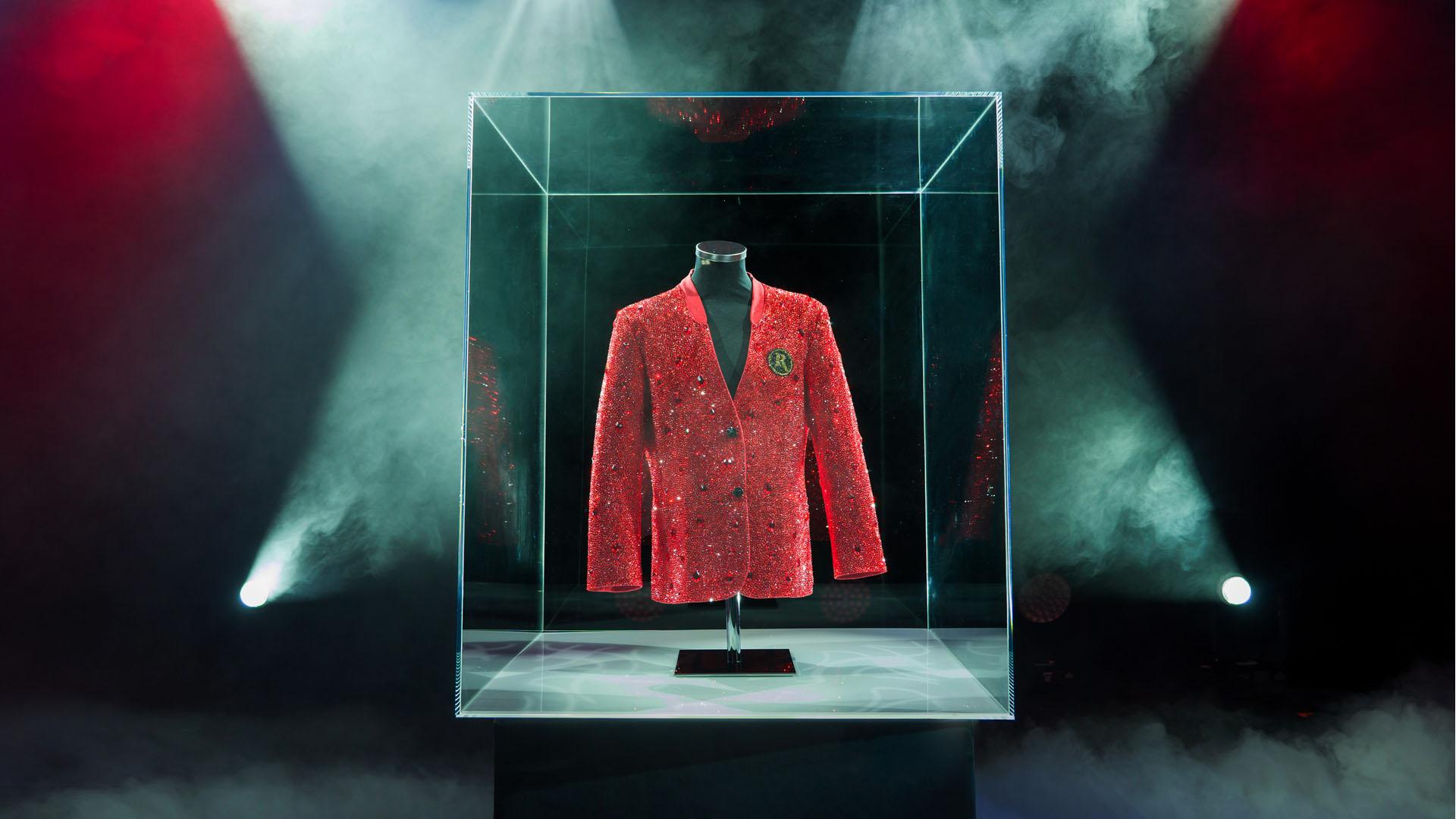 A red rhinestone blazer on a mannequin displayed inside a clear acrylic or glass museum display case on a raised plinth. The case is lit by dramatic spotlights with atmospheric smoke effects in a dark theatrical setting.