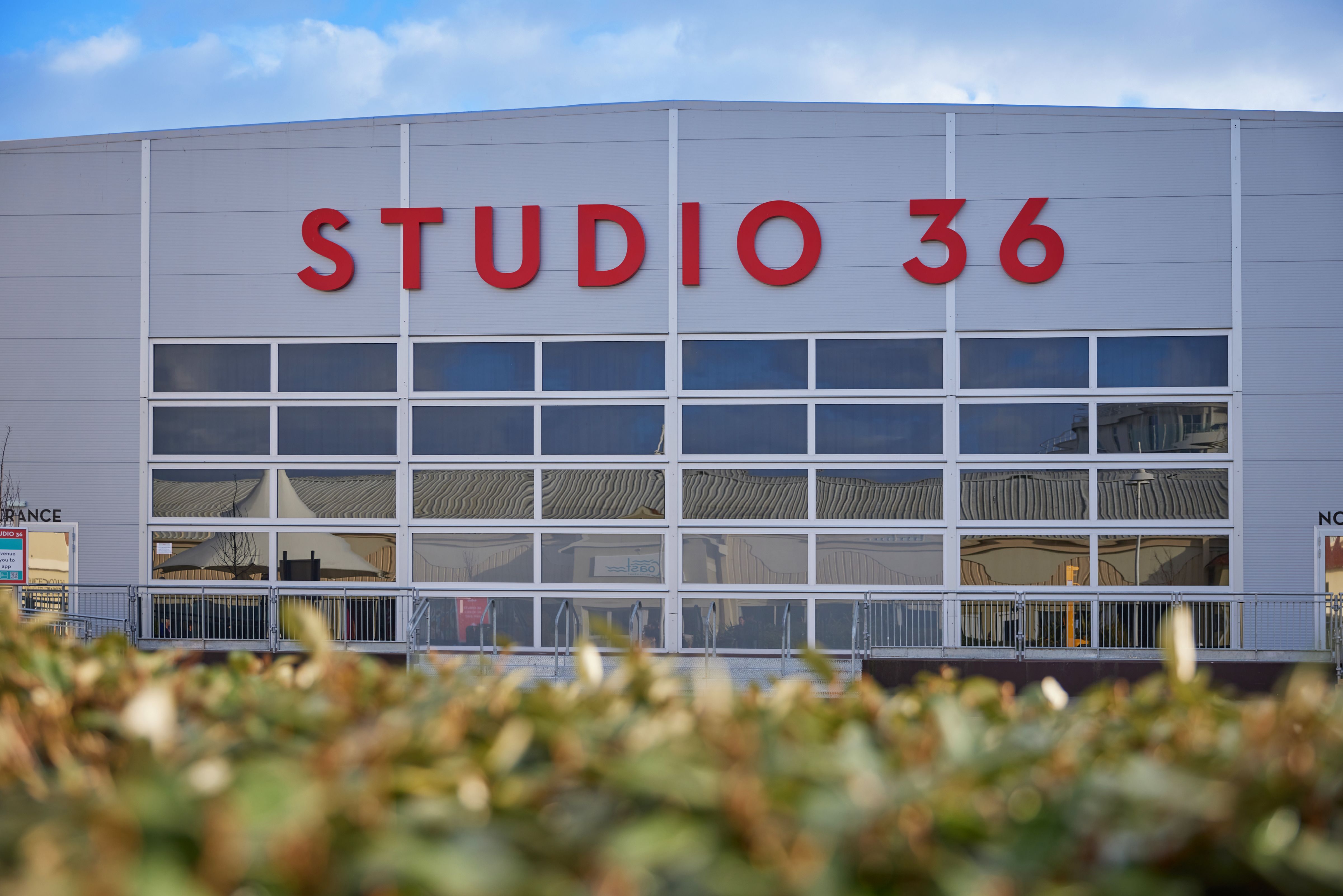 A front-facing view of Studio 36 at Butlin’s, with bold red lettering above large glass panel windows and a hedge in the foreground.