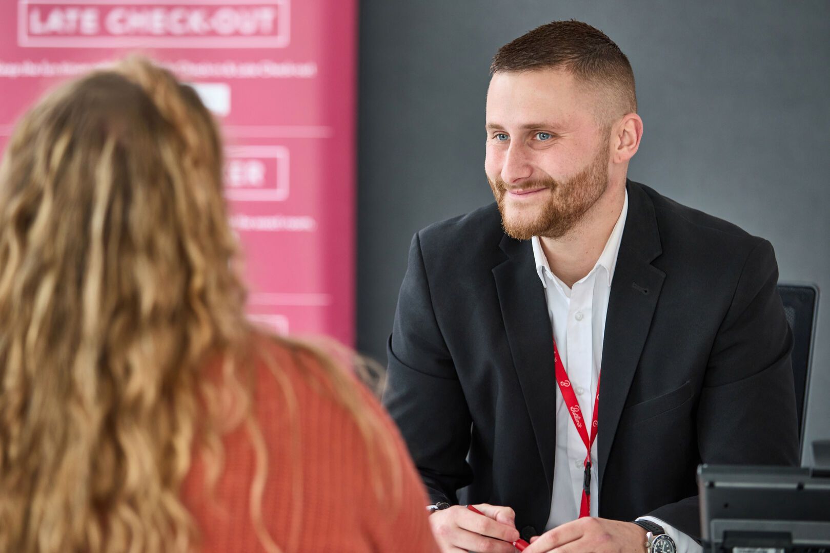 A smiling male Butlin’s team member in a black blazer and white shirt sits at a desk, chatting with a female guest whose back is to the camera. He wears a red Butlin’s lanyard, and a pink sign behind him promotes late checkout options.