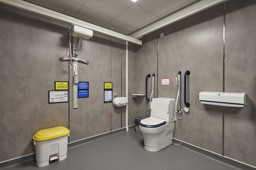 View of the Changing Places facility from the right-hand side, showing a toilet with support rails, accessible paper towel dispensers, and ceiling-mounted hoist system extending across the room.
