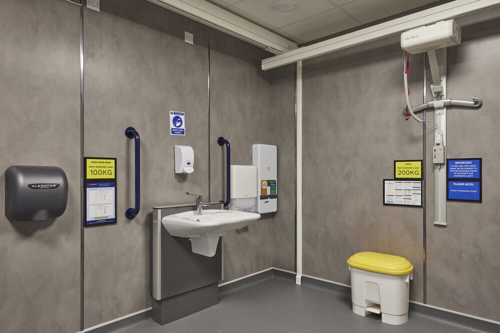 Washbasin area within the Changing Places facility, featuring a sink, soap and hand sanitiser dispensers, hand dryer, safety grab rails, and signage noting maximum working loads.