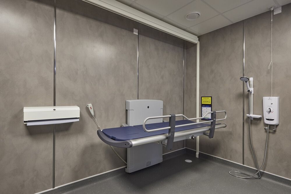 Wide view of a height-adjustable adult changing bench, located next to a wall-mounted shower unit and hand controls. The space is designed for full accessibility.
