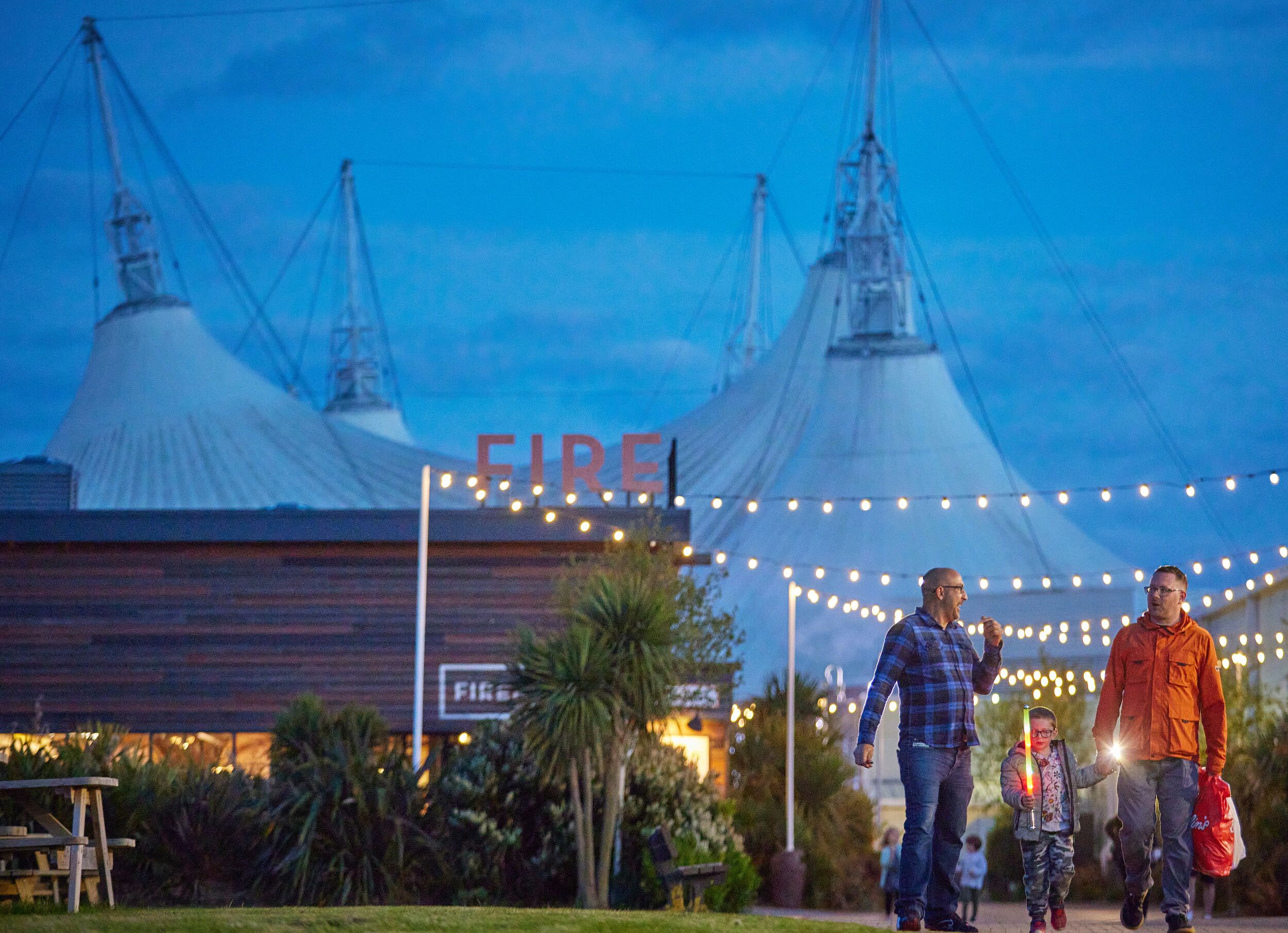 Two adults and a child walking beneath string lights outside the Firehouse Grill at Butlin’s, with the Skyline Pavilion visible in the evening sky.
