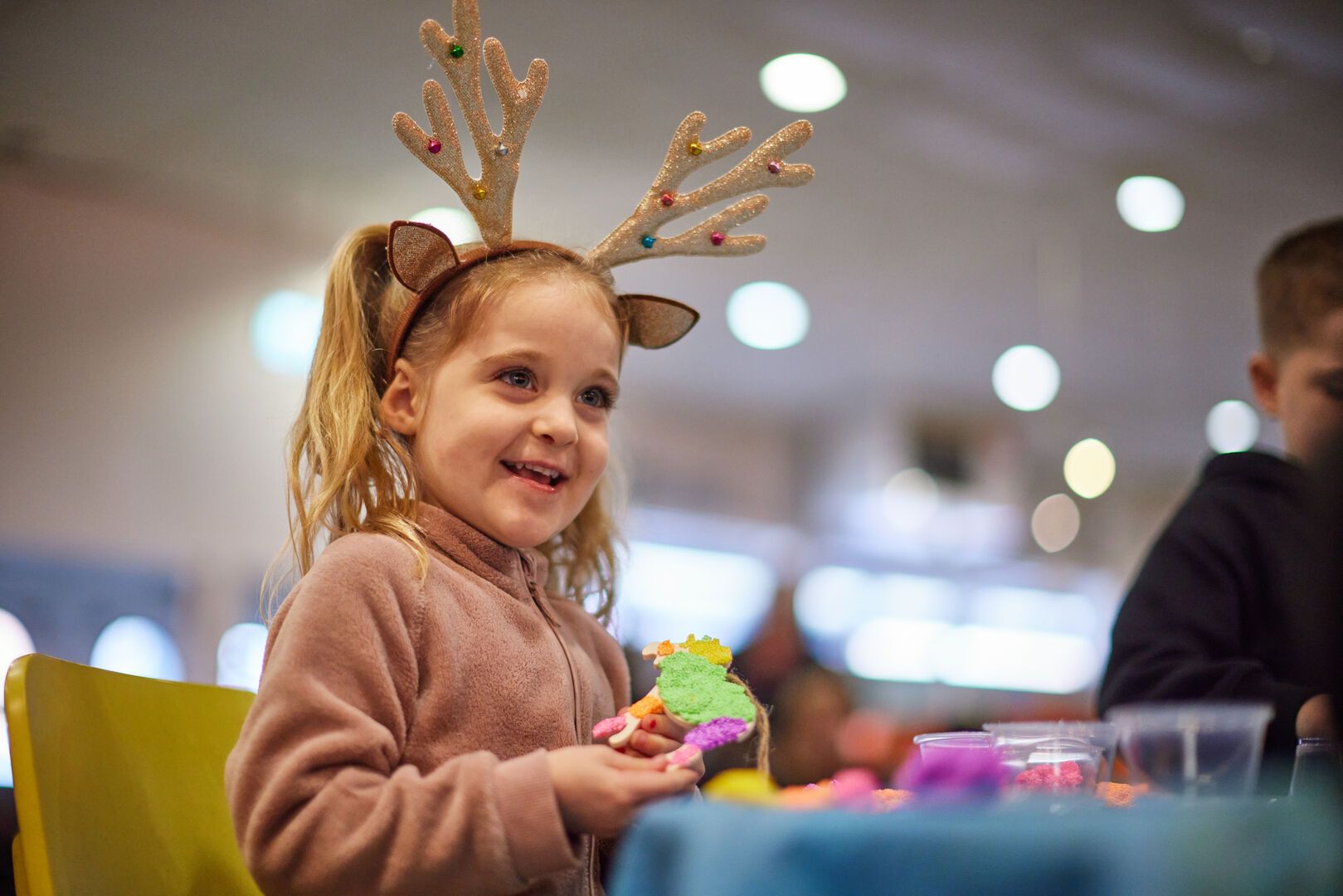 Smiling young girl wearing glittery reindeer antlers holds a colourful handmade decoration during Butlin’s Christmas arts and crafts session.