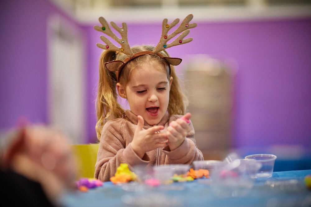 Girl with glittery antlers claps her hands in delight while making Christmas decorations at Butlin’s arts and crafts session, surrounded by colourful materials.