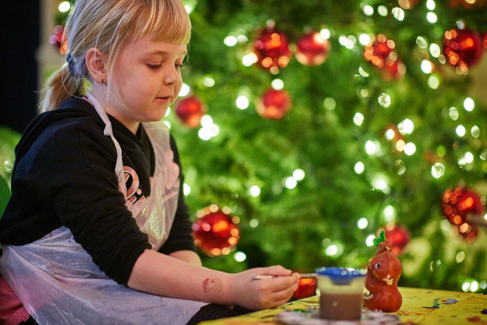 A young girls paints a piece of pottery, with a Christmas tree behind her.