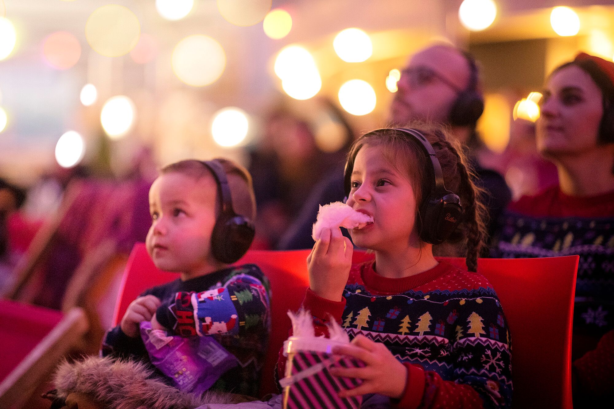 Two young children in festive Christmas jumpers sit side-by-side at Butlin’s Silent Cinema, wearing headphones and snacking on sweets, with warm fairy lights glowing in the background.