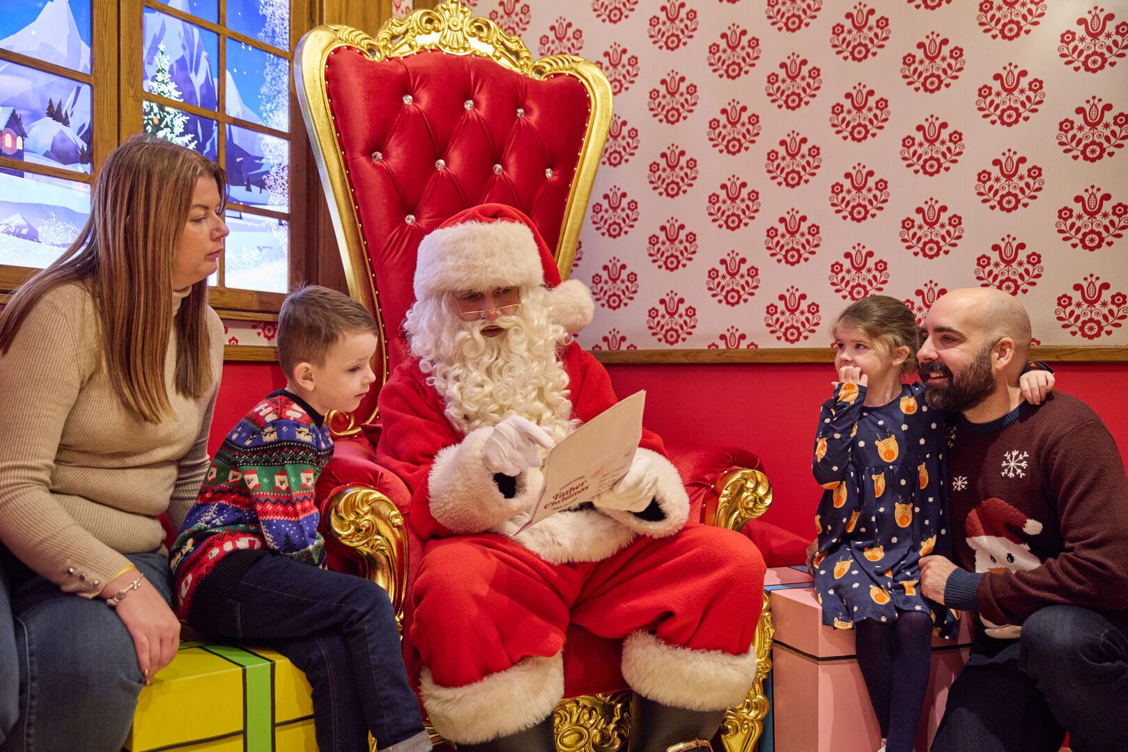 A festive family moment with Santa Claus seated and holding a list and chatting to two children, flanked by smiling parents. The setting is a warmly lit, wintry room with patterned red wallpaper and snowy outdoor scenes visible through the window.