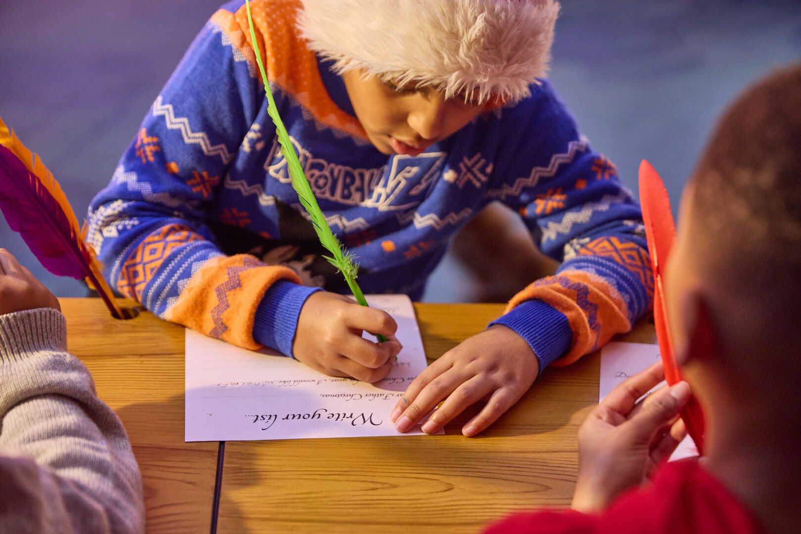 A child in a festive jumper and Santa hat concentrates on writing their Christmas list using a green feather quill. They sit at a wooden table next to others holding colourful feathers.