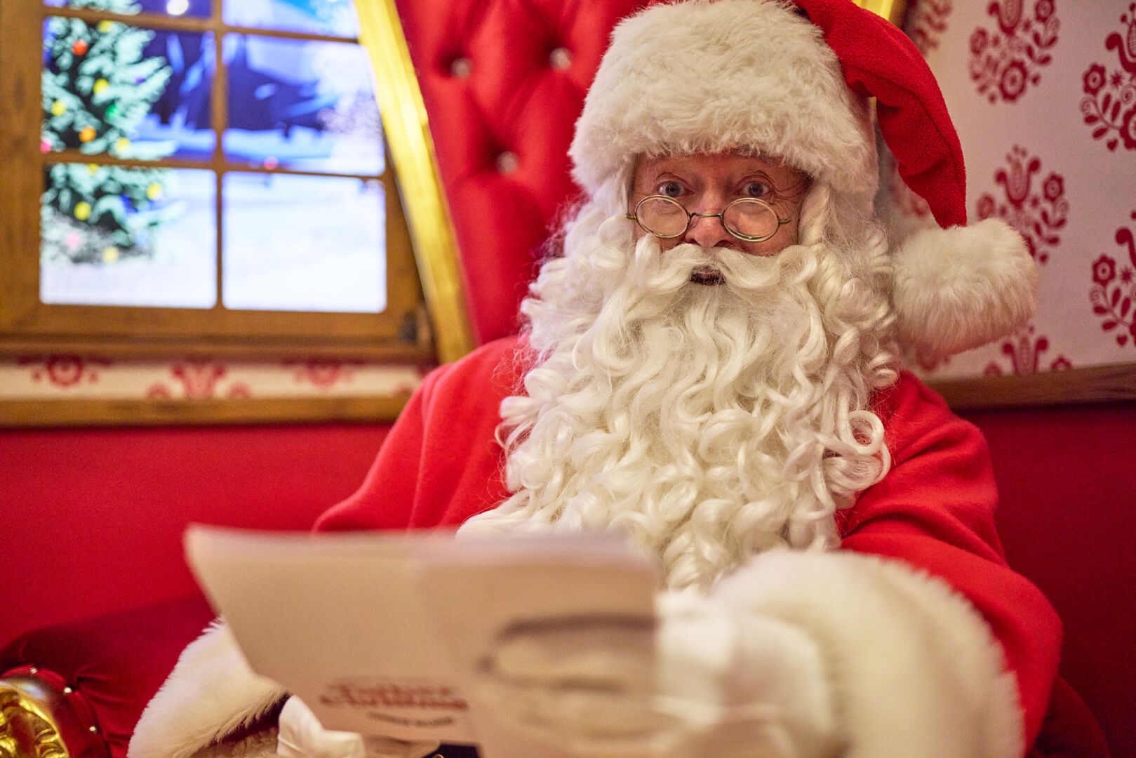 Father Christmas sits alone in his grotto reading a festive wish list. He’s framed by a large golden throne, plush red bean bags, and a digital snowy backdrop through the window, creating a quiet, magical moment before greeting the next family.