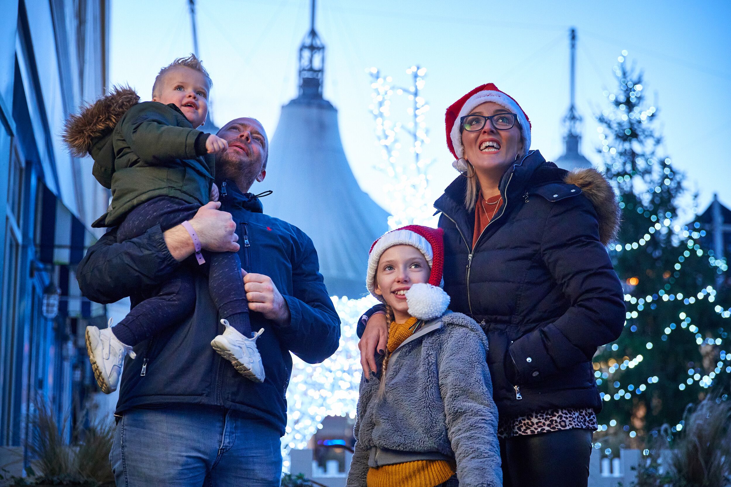 Family of four wearing winter coats and Santa hats admiring festive Christmas lights at Butlin’s resort.
