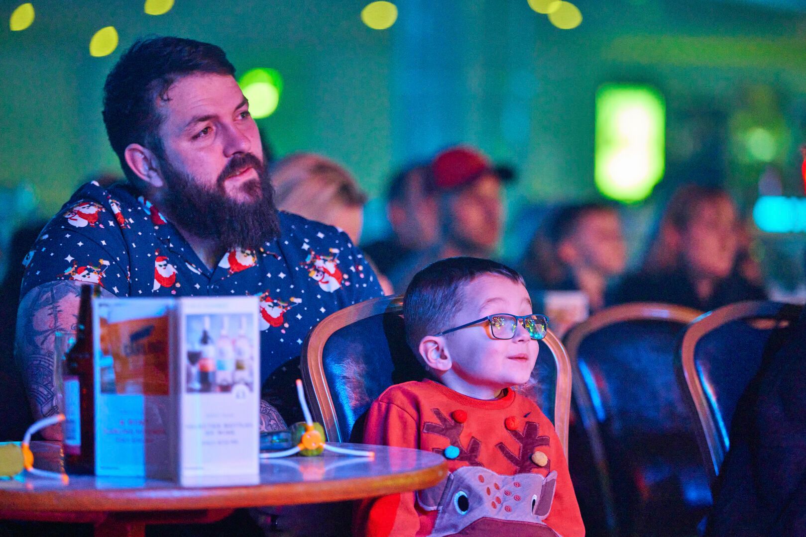 A father and son dressed in festive clothing, sit together at a table while watching a show.