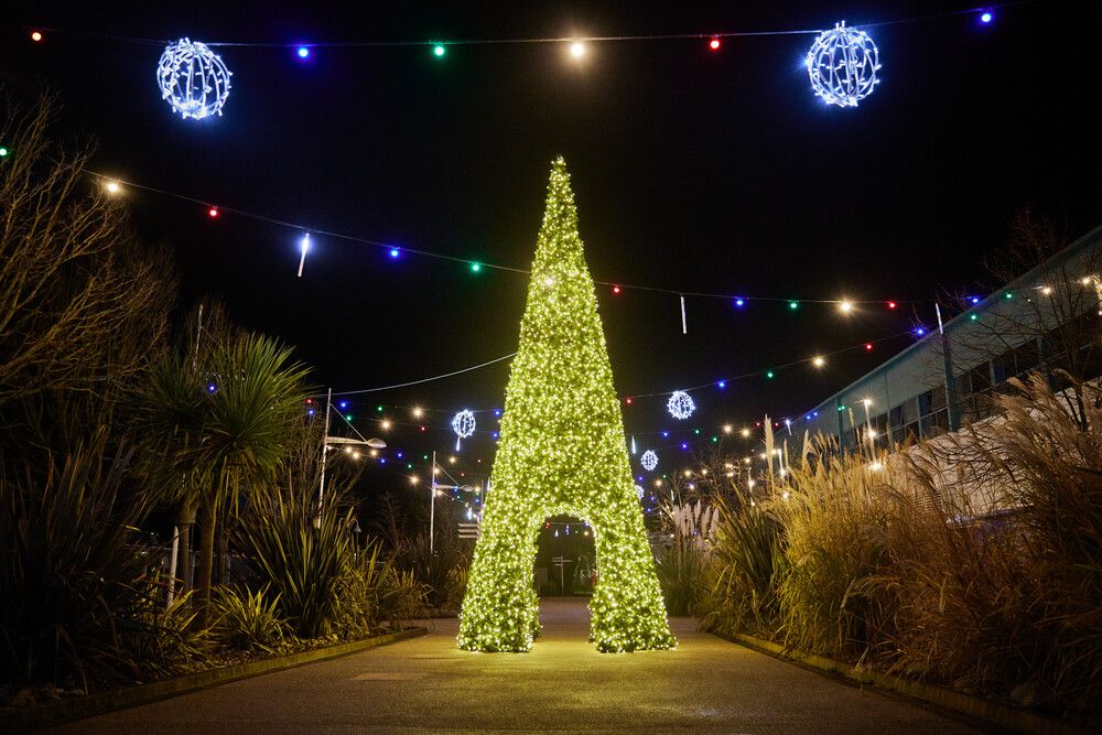 A tall, cone-shaped Christmas tree covered in green fairy lights forms an illuminated archway along a festive outdoor path. The surrounding area is decorated with hanging multicoloured string lights and glowing snowflake-shaped lights.