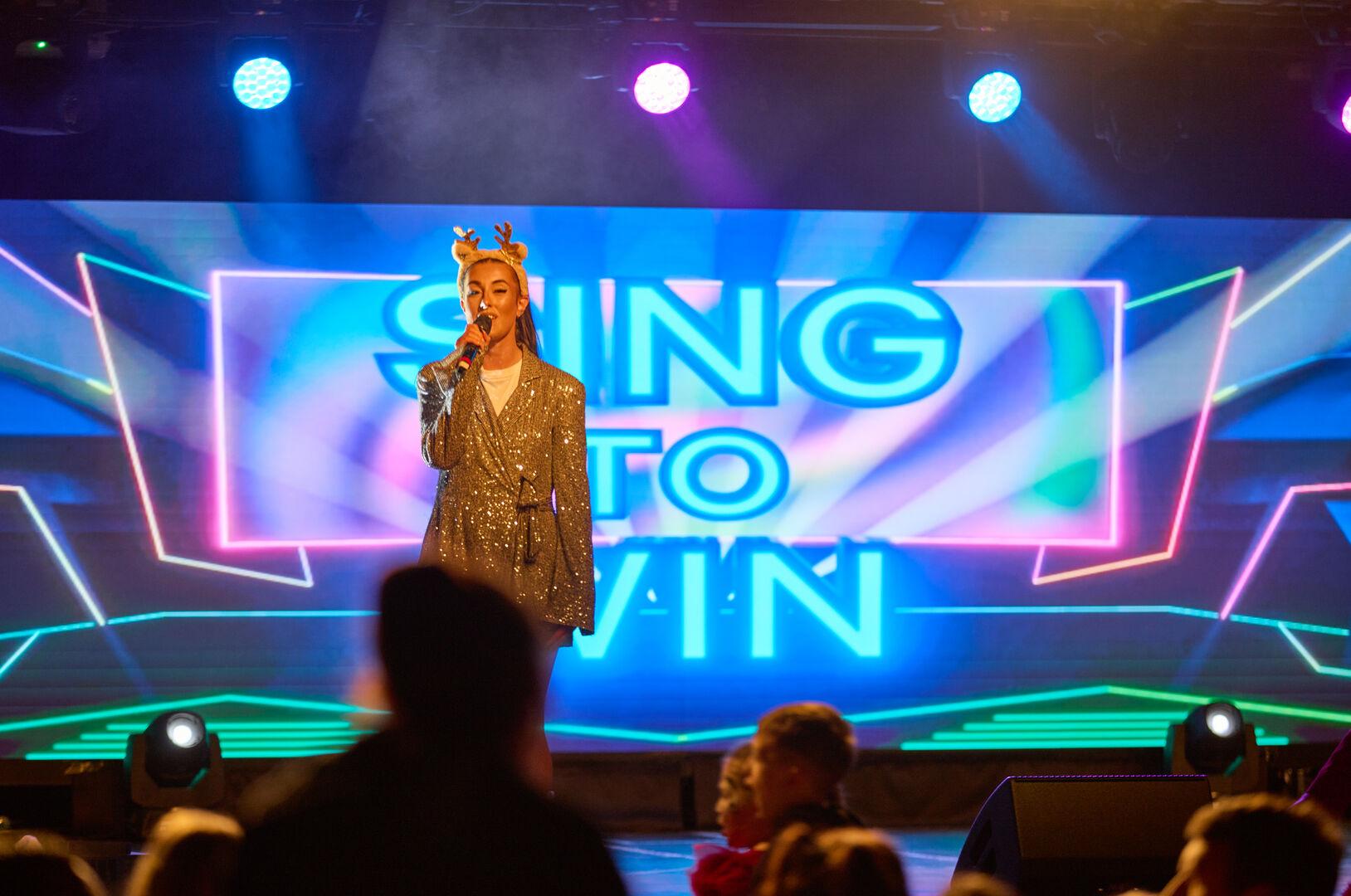 A performer in sparkly silver attire stands on stage singing into a microphone, framed by a digital screen that reads “SING TO WIN” in bright neon letters. She wears reindeer antlers, adding a festive touch to the scene. Children are gathered at the front of the stage, lit by warm lighting.