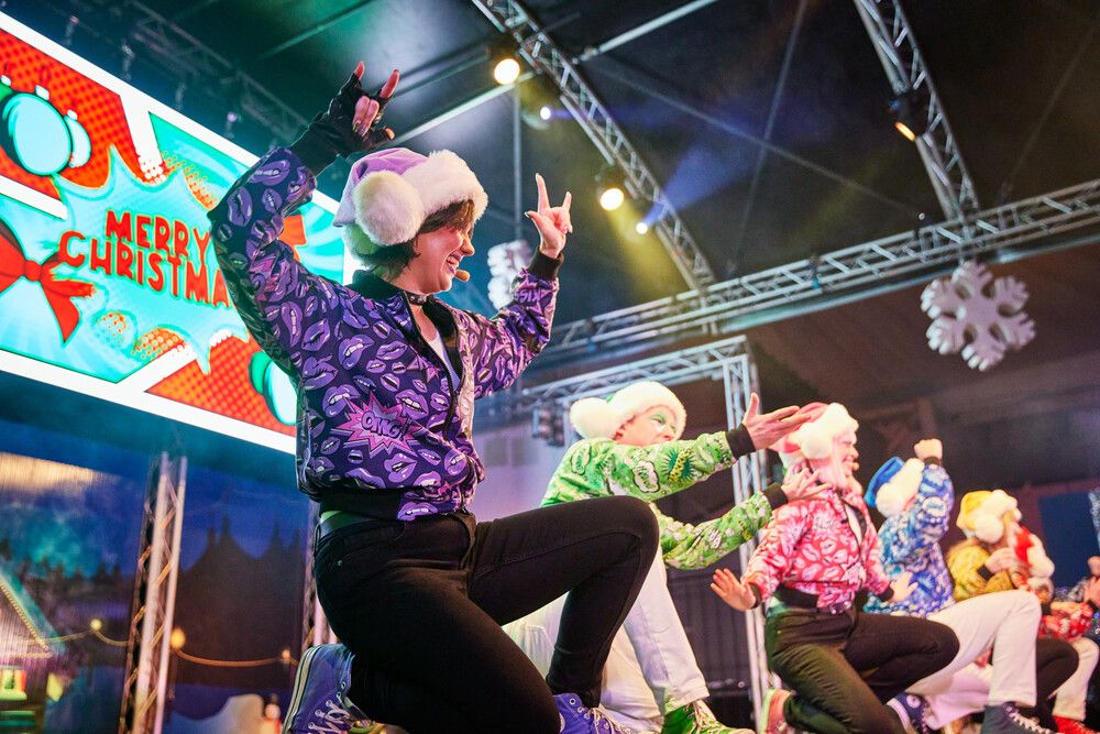 A dynamic stage moment during the Skyline Gang's Christmas Goes Pop show, with cast members kneeling in a row mid-dance, all wearing comic-print jackets and Santa hats. One performer in a purple jacket and white gloves leans forward, striking a rock-and-roll pose. The colourful “Merry Christmas” comic-style screen glows in the background.