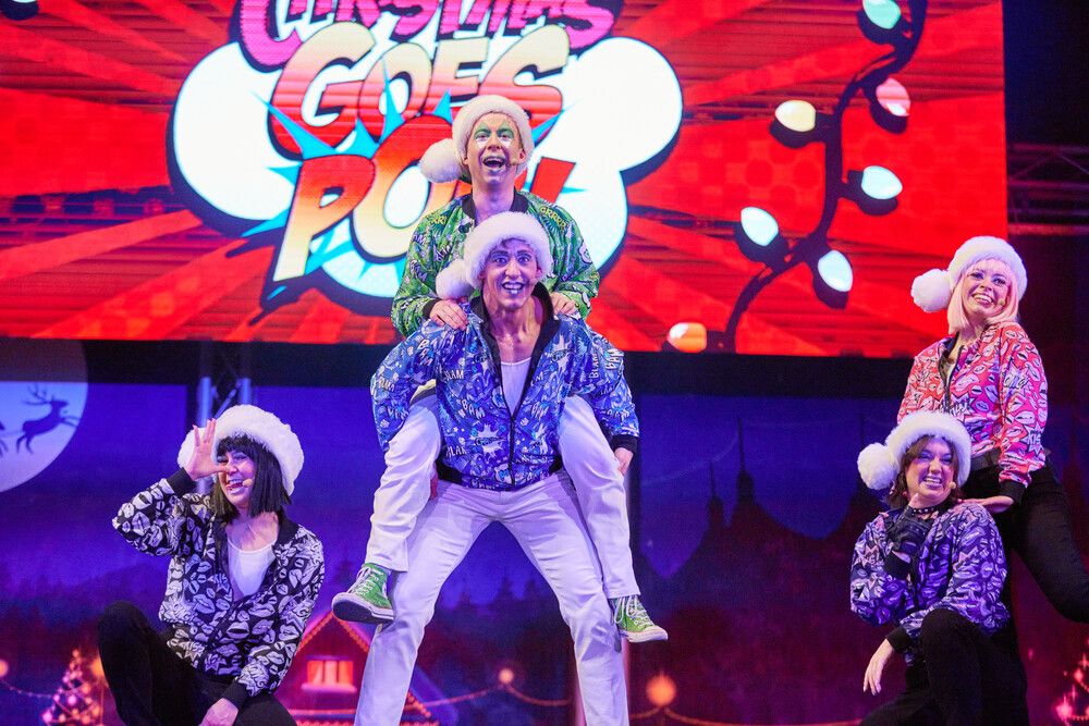 The Skyline Gang on stage during a festive performance, with one performer in a green jacket sitting on the shoulders of another in a blue jacket. All five characters wear Christmas hats and colourful patterned jackets, with the "Christmas Goes Pop!" logo brightly displayed behind them on a screen.