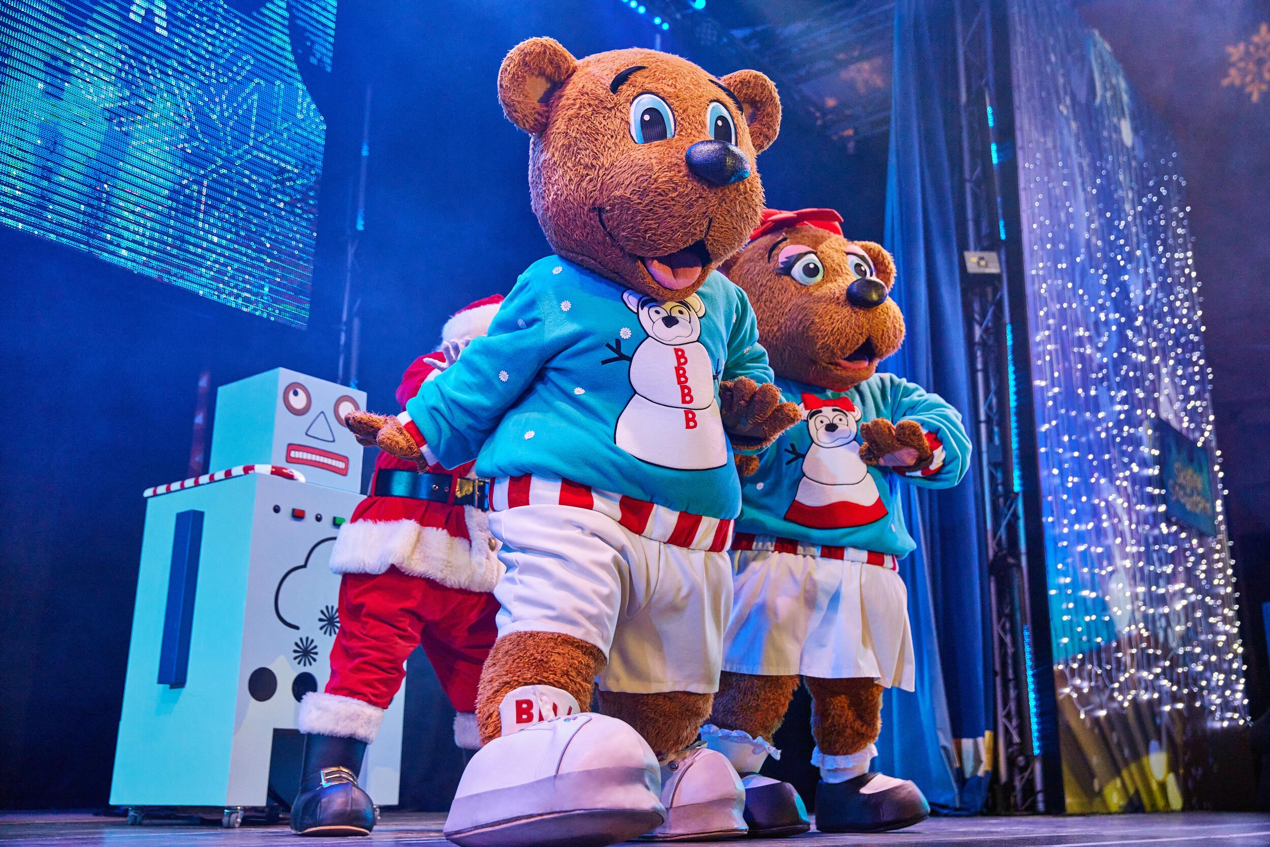 A cheerful Billie Bear is front and centre, mid-performance with Santa and Bonnie Bear just behind. Billie wears a turquoise snowman jumper and festive shorts, while Bonnie sports her iconic bow and skirt. A crowd of families watches from below as snow falls and music plays.