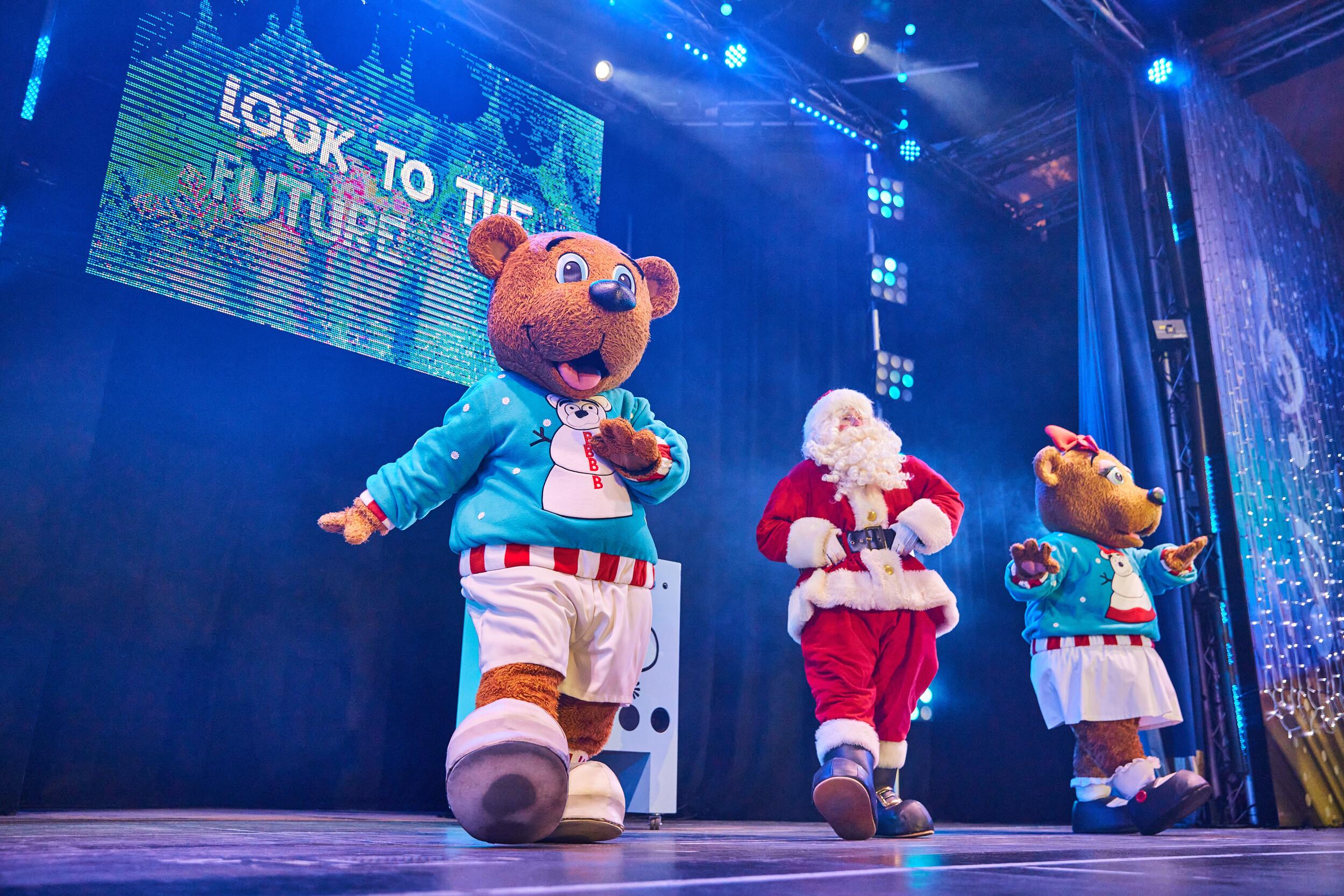 Billie Bear leads a fun-filled dance routine with Santa and Bonnie Bear during the Skyline Snowstorm show. All three characters are in motion with enthusiastic expressions under a screen that reads “LOOK TO THE FUTURE.” Blue and purple lighting and festive sparkle curtains create a vibrant, magical atmosphere.
