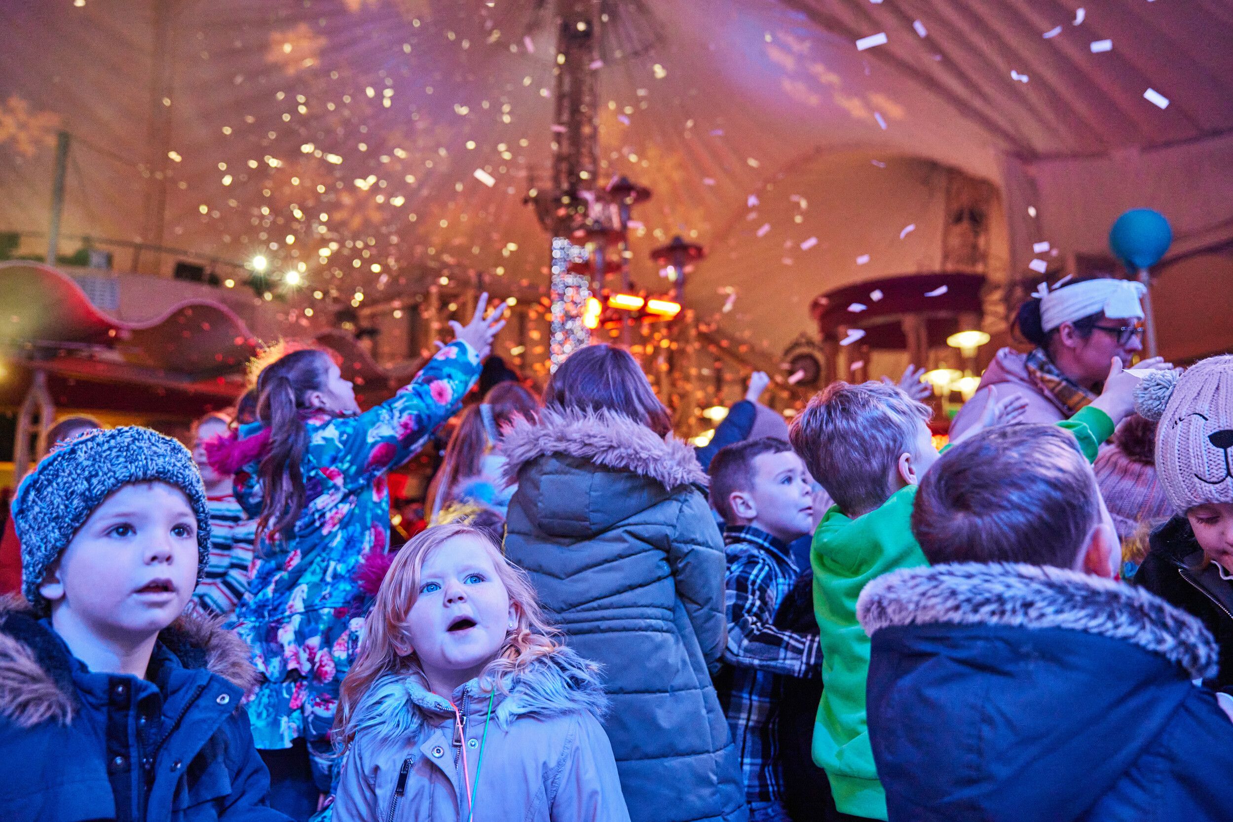 A crowd of warmly dressed children watch in awe as artificial snow falls inside the Skyline Pavilion. Confetti catches the colourful lights, while a girl in a floral coat reaches into the air.