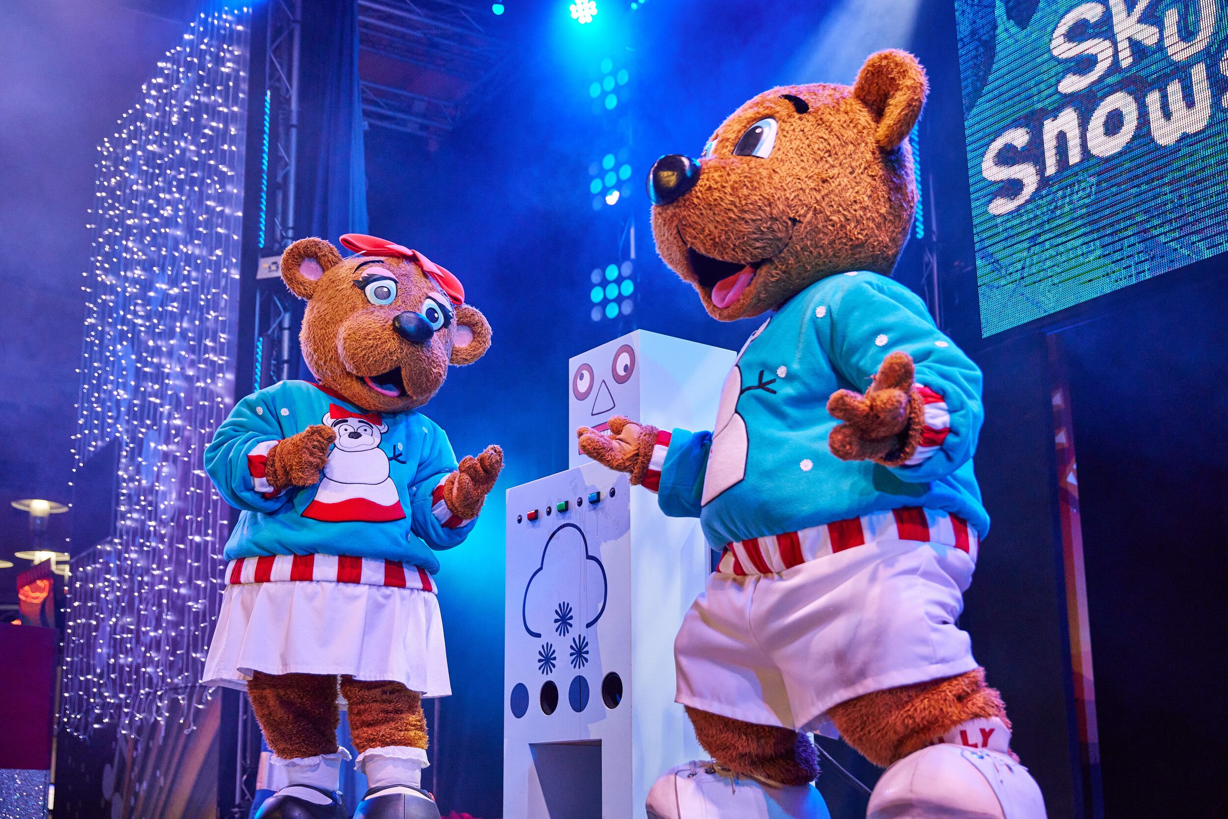Billie and Bonnie Bear interact beside a whimsical control panel decorated with snowflake and weather symbols. Both are dressed in matching turquoise festive jumpers with cartoon snowmen. Bonnie looks intrigued while Billie gestures animatedly, lit by a blue stage spotlight.