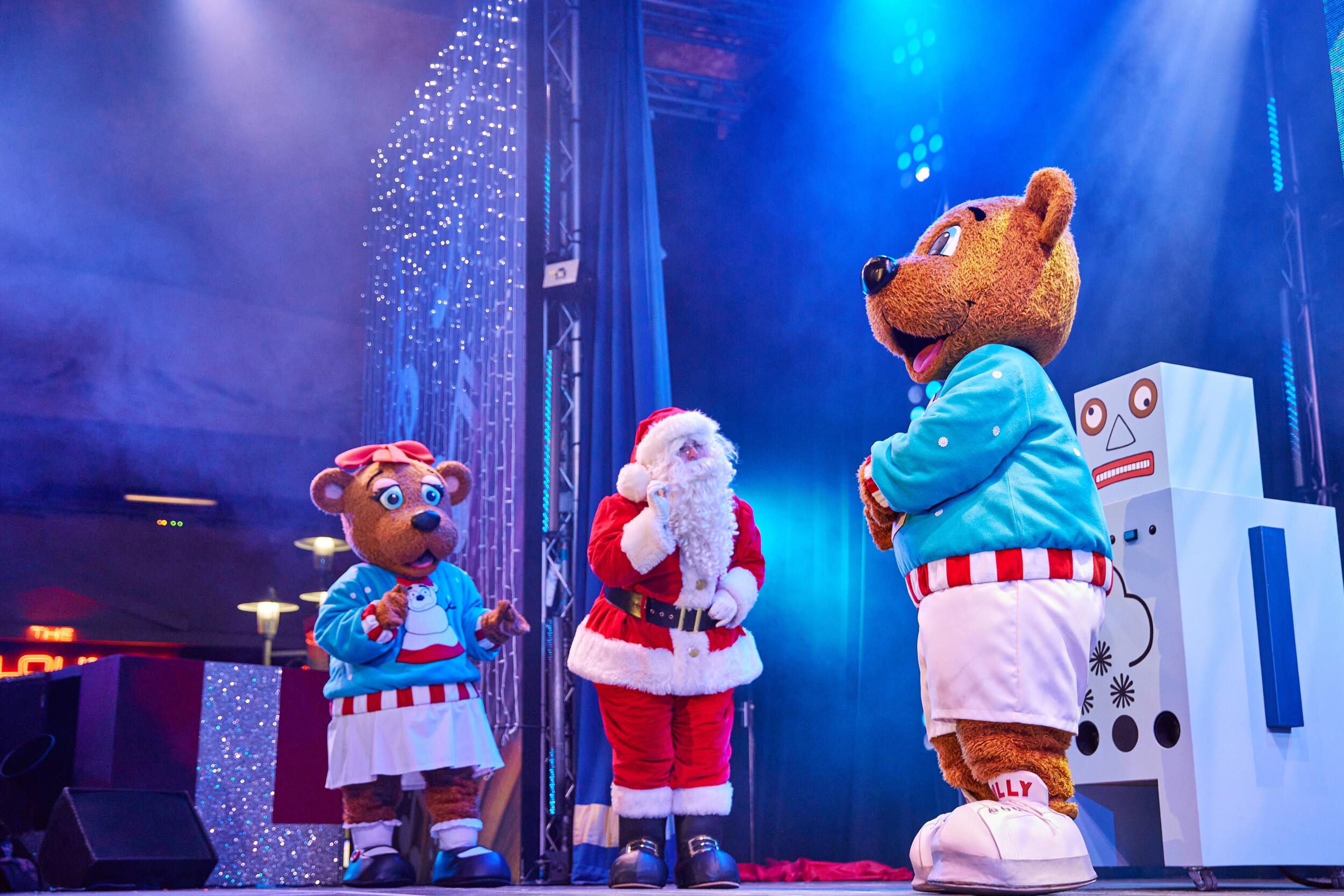 Santa Claus stands between Billie and Bonnie Bear, all three characters seemingly mid-conversation during the Skyline Snowstorm show. Behind them is a quirky snow machine and strings of sparkling lights. Santa strokes his beard thoughtfully while the bears listen attentively.