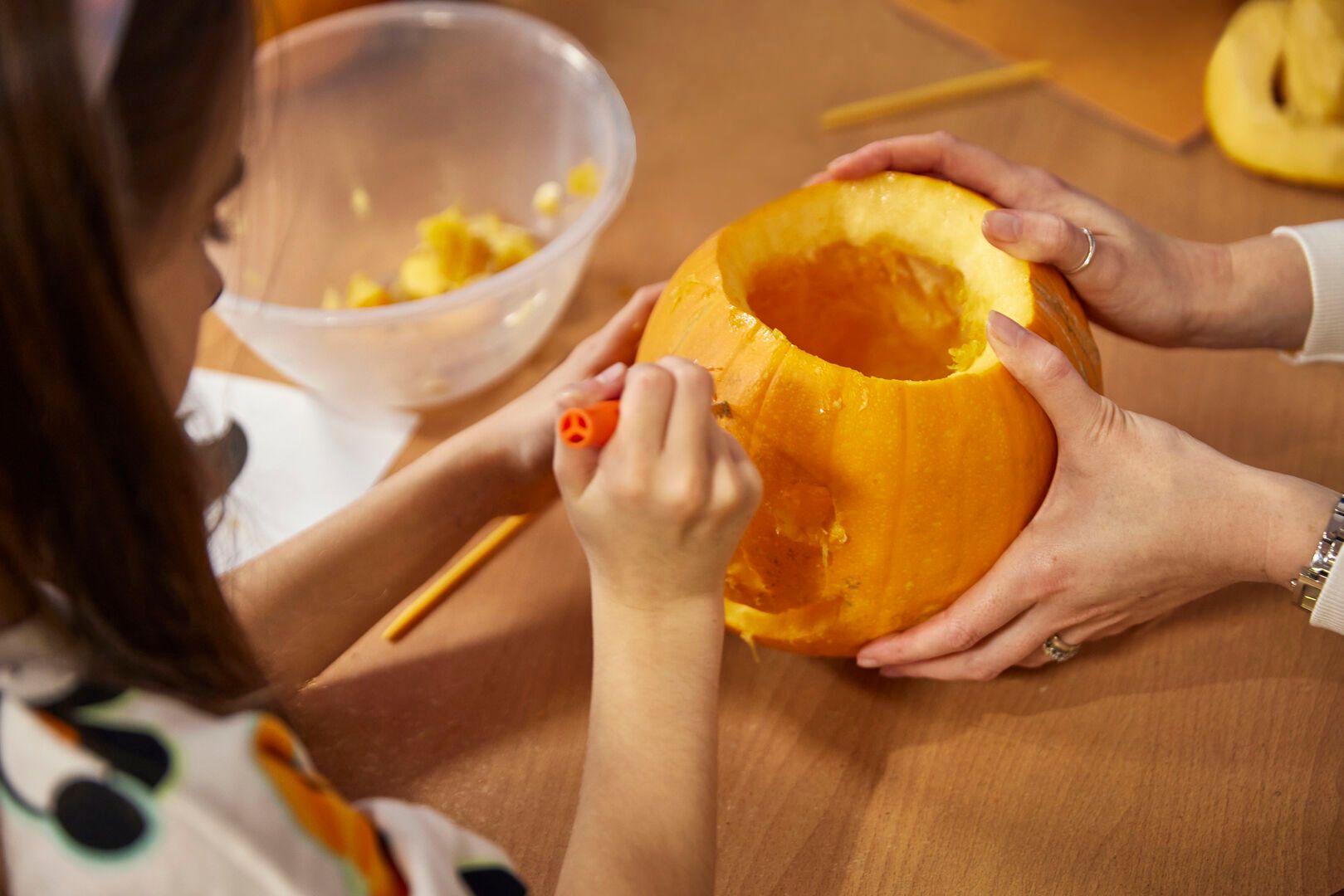 A young girl carves a pumpkin with help from an adult.