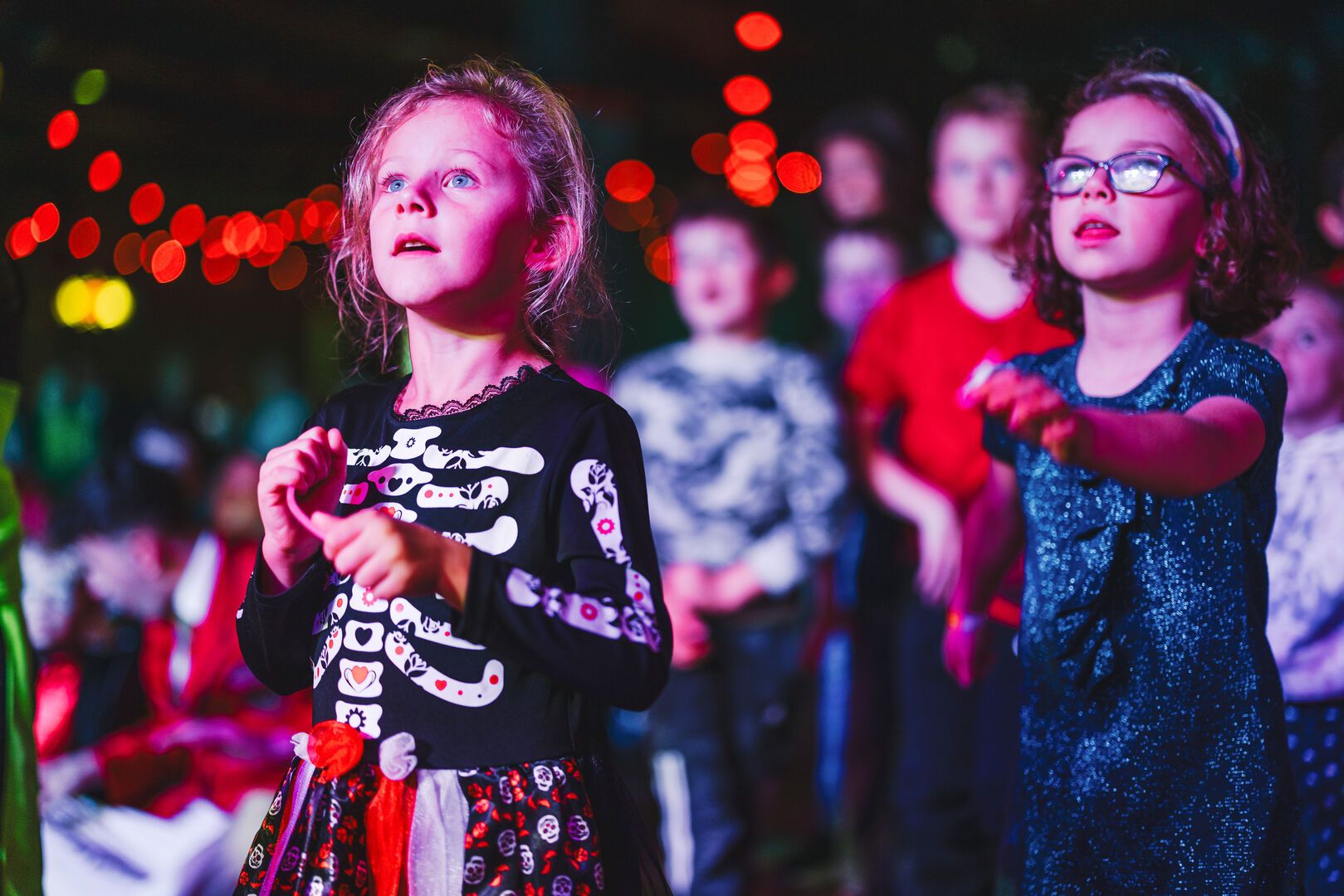 A young girl, dressed in a Skeleton costume, watches a show in a crowd of children.