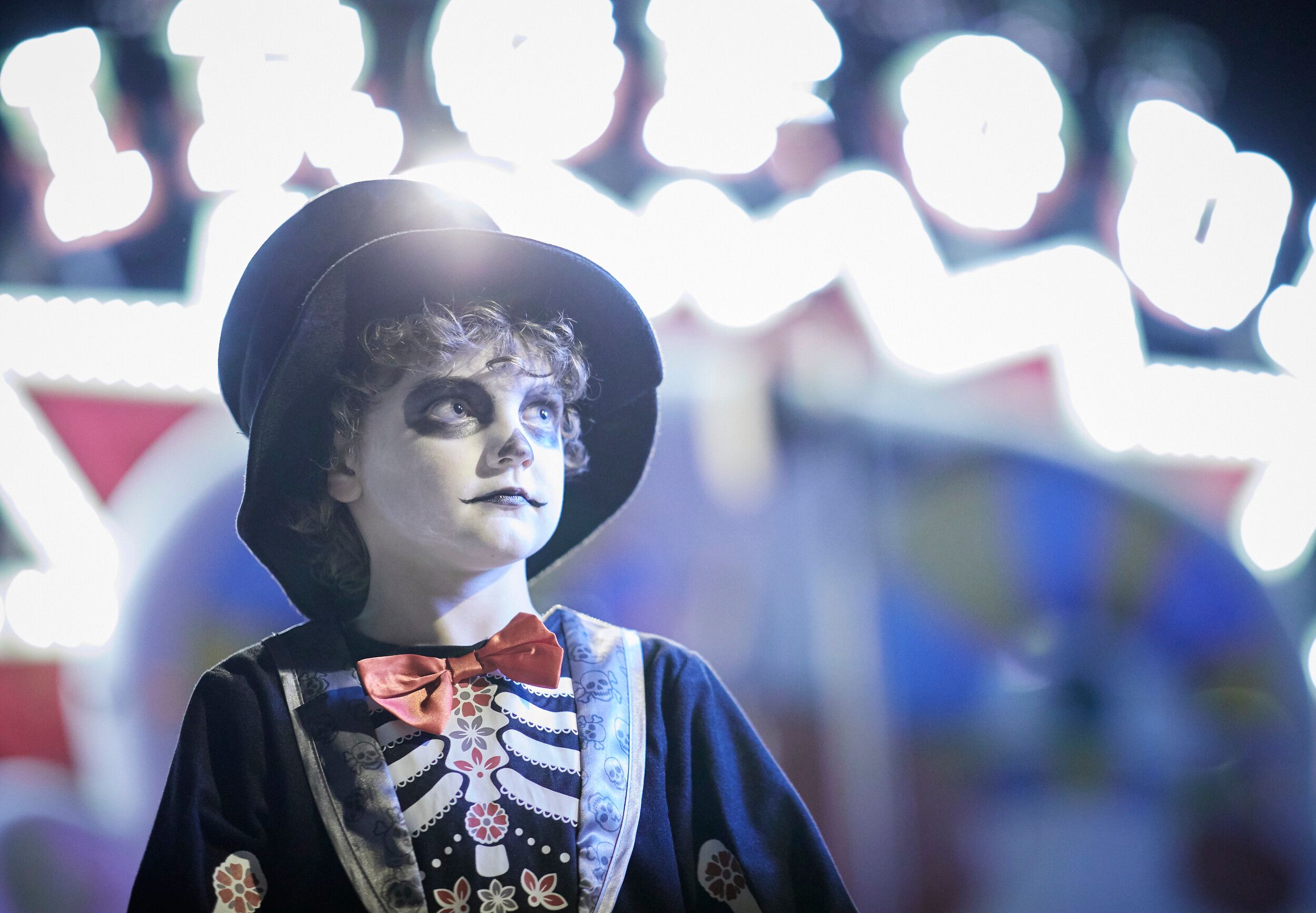 A young boy dressed in a spooky skeleton costume with face paint and a top hat, standing in front of a brightly lit, slightly blurred fairground backdrop at night.