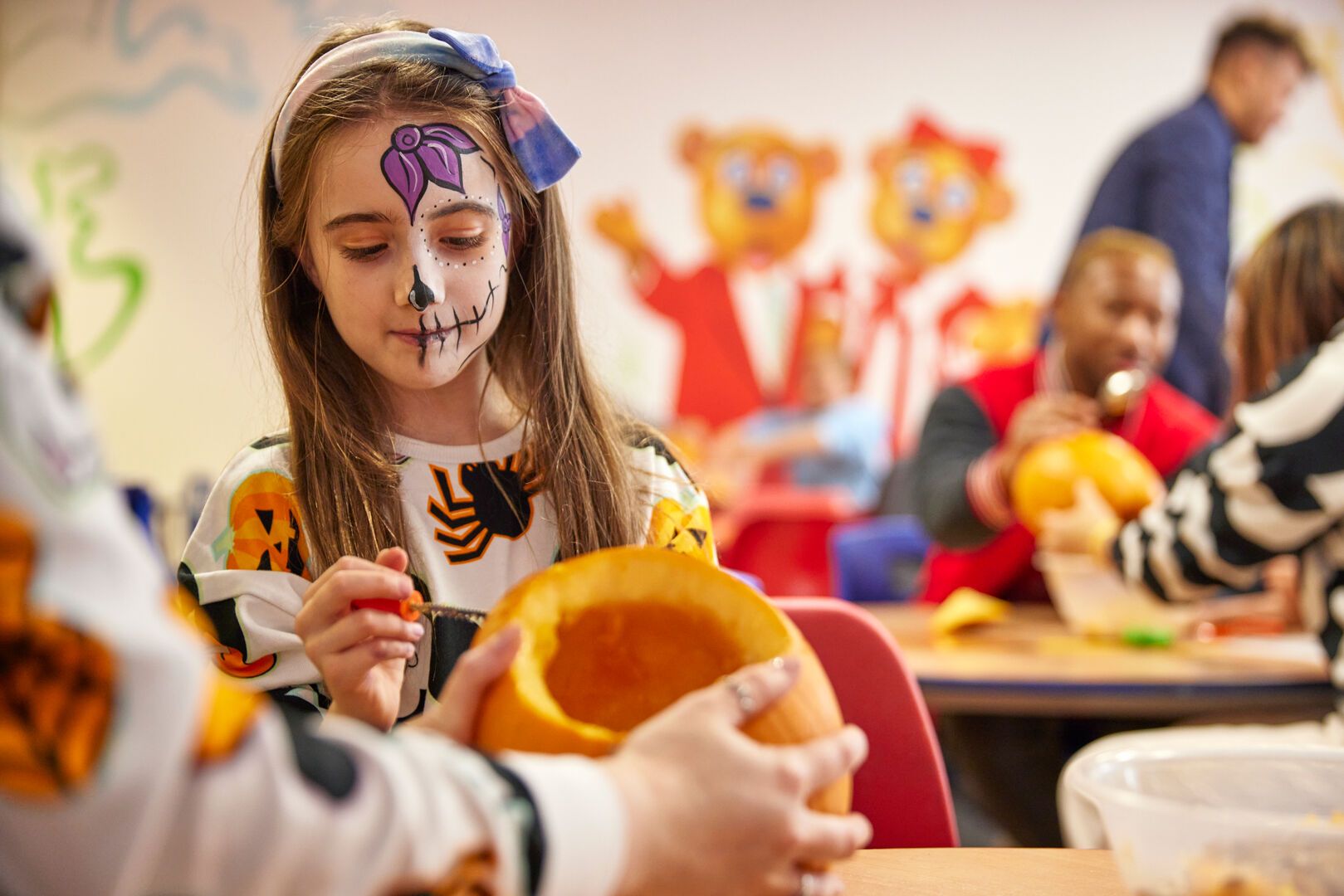 A girl with painted sugar skull face paint carves a pumpkin at a craft table, with colourful decorations and families in the background.