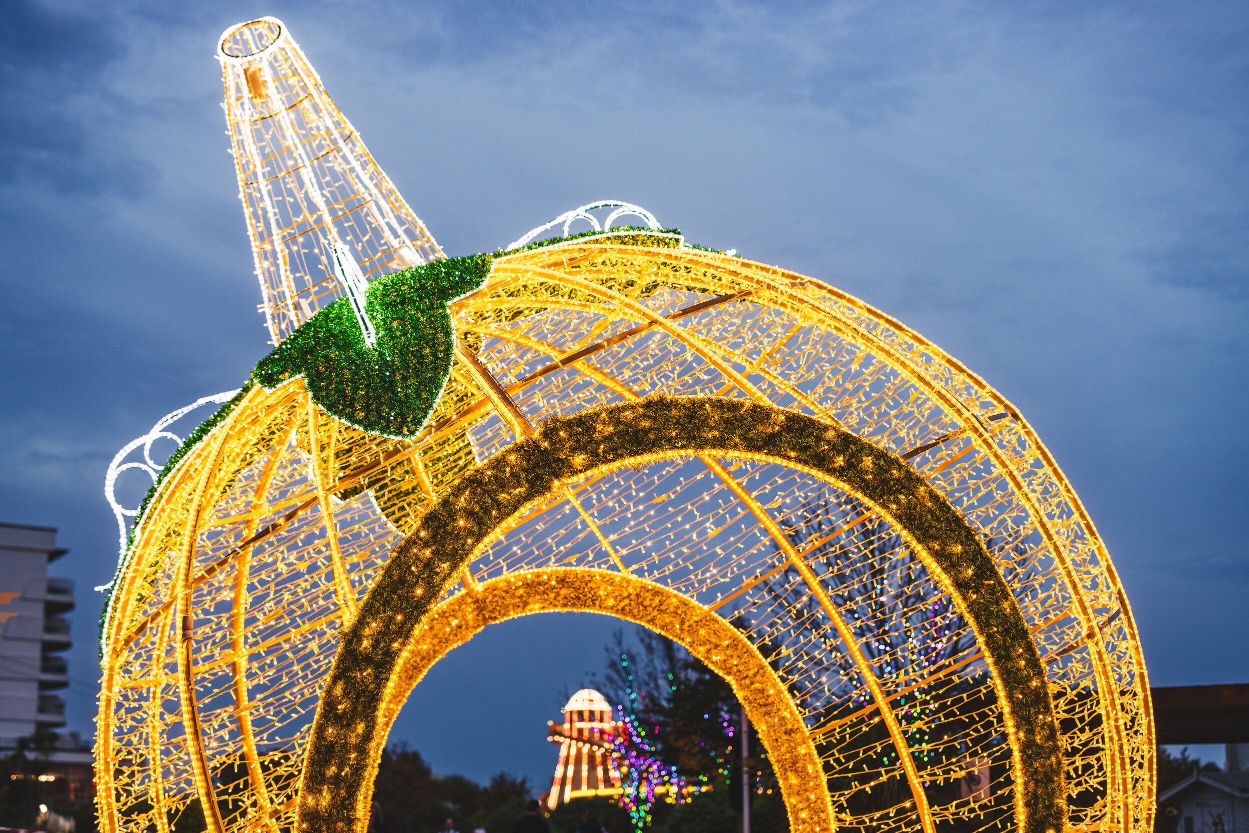 A giant pumpkin-shaped light sculpture outdoors at dusk, glowing gold with a green leaf on top, and more illuminated attractions visible through its arch.