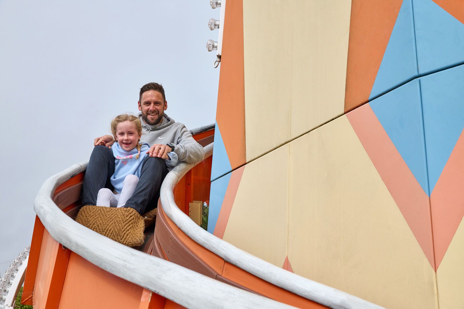A man and young girl ride together on the Butlin’s outdoor helter-skelter, both smiling and seated on a slide mat as they descend the colourful spiral.