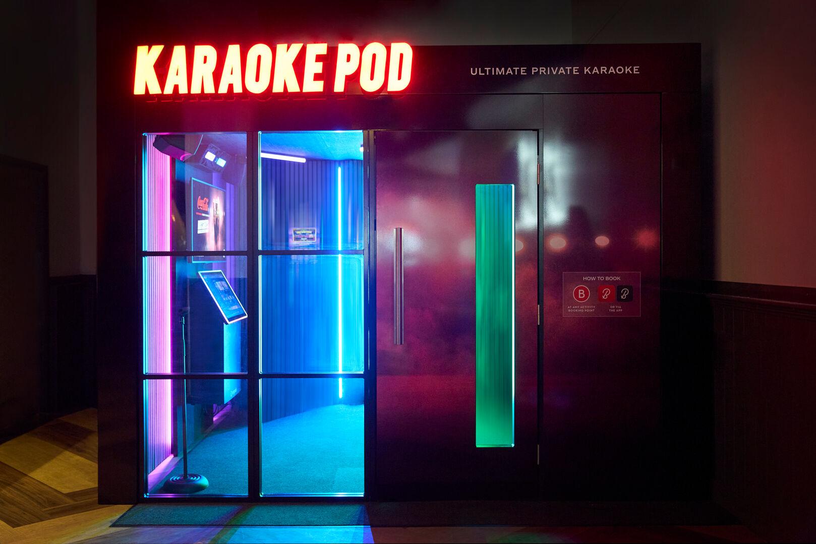 A private karaoke pod booth with a glowing neon sign reading "KARAOKE POD" and the tagline "Ultimate Private Karaoke." The pod features glass-panelled doors illuminated by vibrant pink, blue, and green neon strip lighting. Inside, a microphone stand and tablet screen are visible. A solid dark door on the right side displays booking instructions.