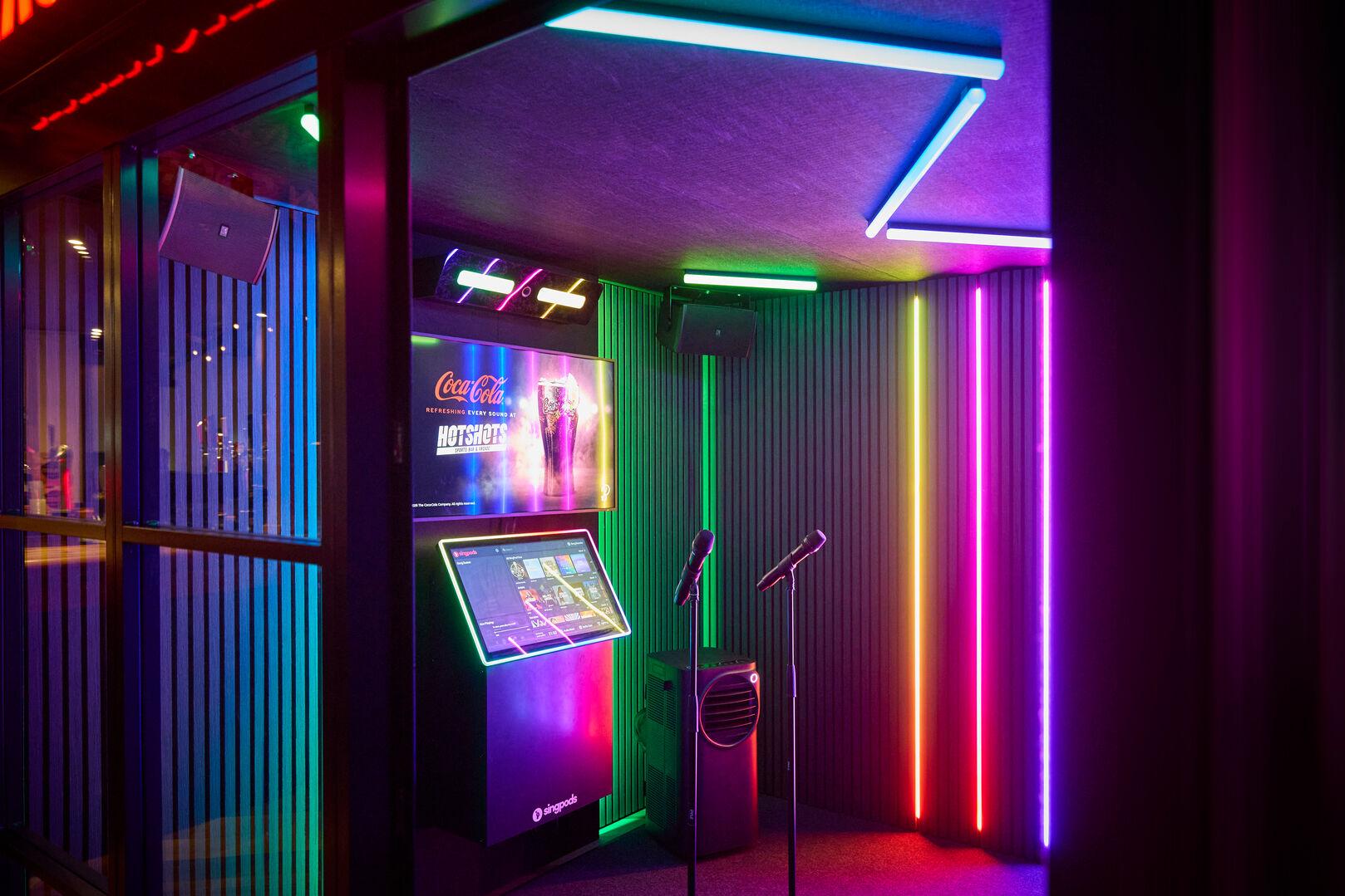 The interior of a private karaoke pod by Singpods, bathed in vibrant rainbow neon lighting across the walls and ceiling. The interior of a private karaoke pod by Singpods, bathed in vibrant rainbow neon lighting across the walls and ceiling. Two microphones on stands are positioned centre stage in front of a large touchscreen song selection kiosk and a wall-mounted screen displaying a Coca-Cola and Hotshots advertisement. Surround sound speakers are mounted on the walls, and acoustic panelling lines the interior for sound quality.