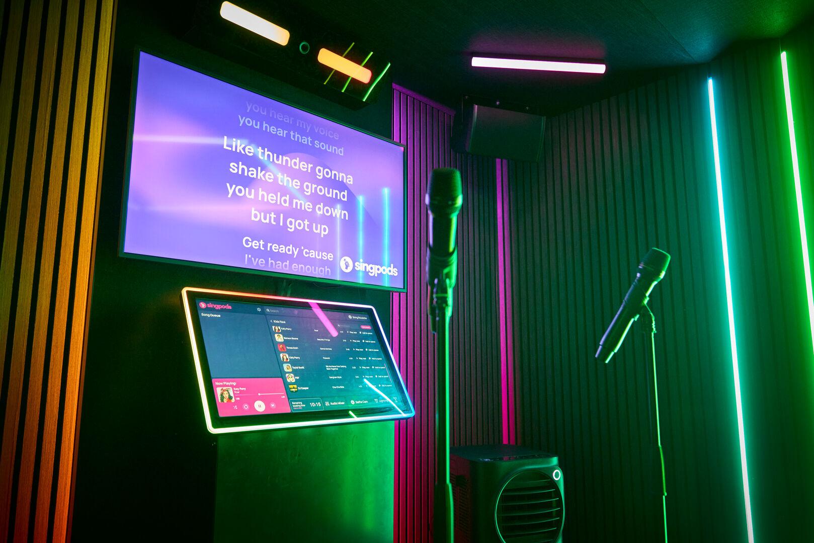 The interior of a Singpods karaoke pod mid-session, with lyrics displayed on a wall-mounted screen against a purple gradient background. The interior of a Singpods karaoke pod mid-session, with lyrics displayed on a wall-mounted screen against a purple gradient background. A touchscreen kiosk below shows the song queue interface with artists including Katy Perry and Taylor Swift. Two microphones on stands are positioned in the foreground, with green and pink neon strip lighting illuminating the acoustic-panelled walls.