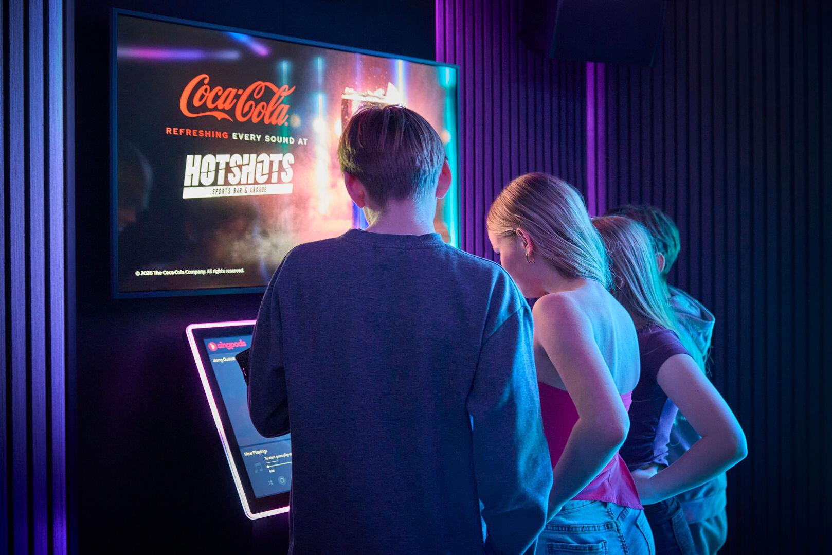 Three young people seen from behind browsing the Singpods song selection kiosk inside a karaoke pod. A large wall-mounted screen above displays a Coca-Cola advertisement for Hotshots Sports Bar and Arcade. The pod interior is lit with purple and blue neon lighting against dark acoustic-panelled walls.