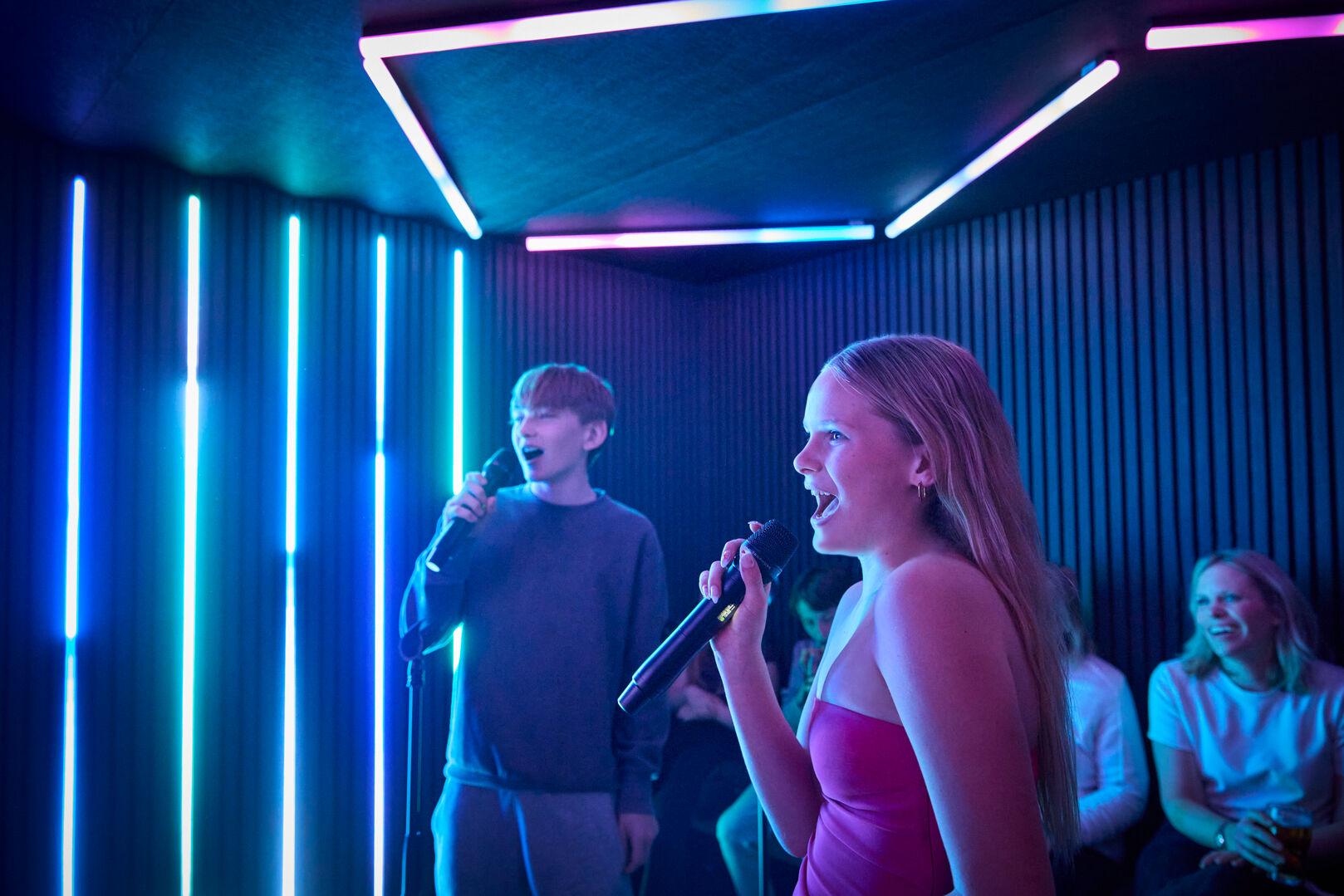Two young people singing into microphones inside a karaoke pod, both mid-performance with mouths open. A young woman in a pink dress holds her microphone in the foreground, while a young man in a grey sweatshirt sings in the background. Friends can be seen seated and laughing behind them, with tall blue and white neon strip lights lining the acoustic-panelled walls and ceiling.