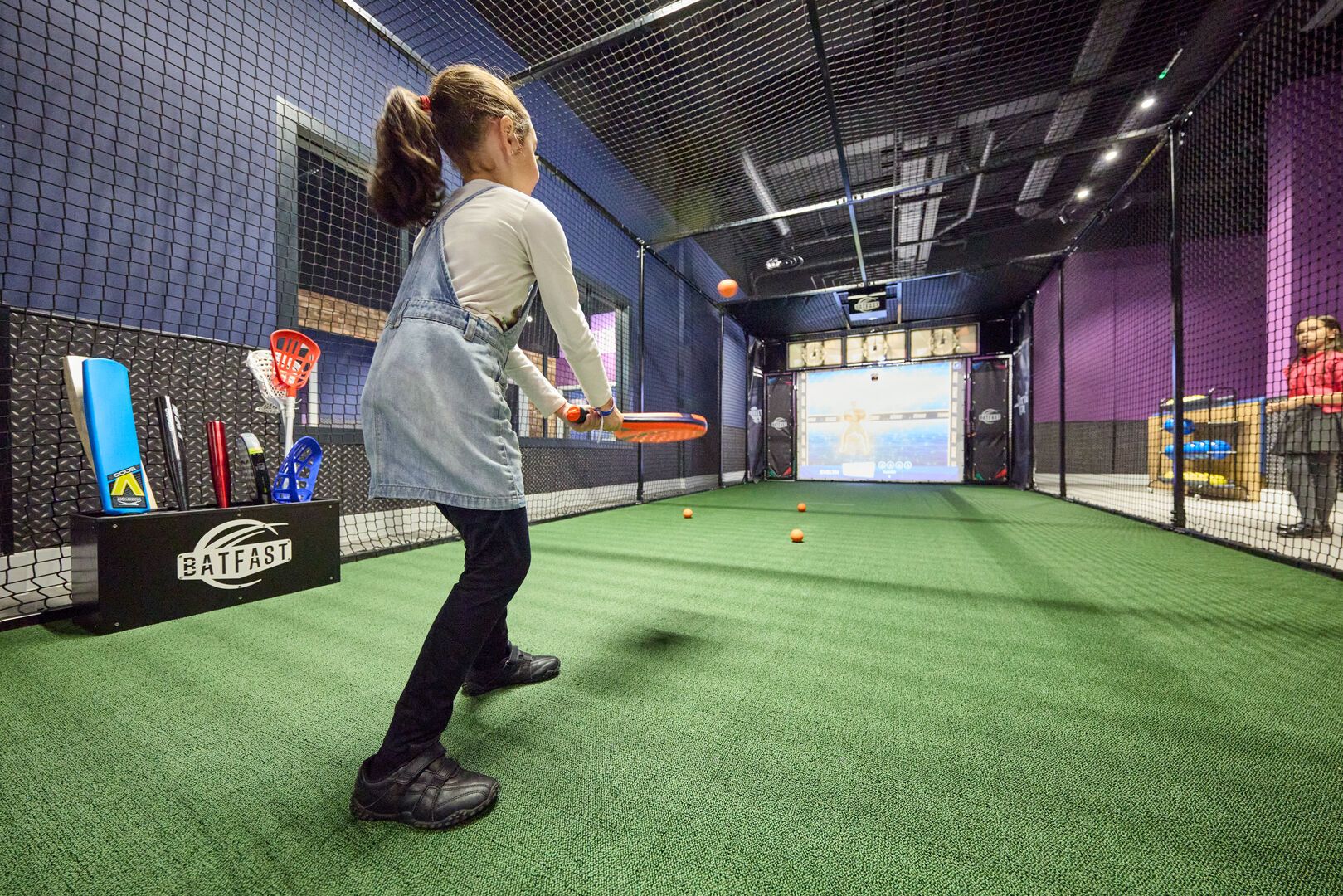A young girl playing a game at the Batting Cage with a tennis racket.