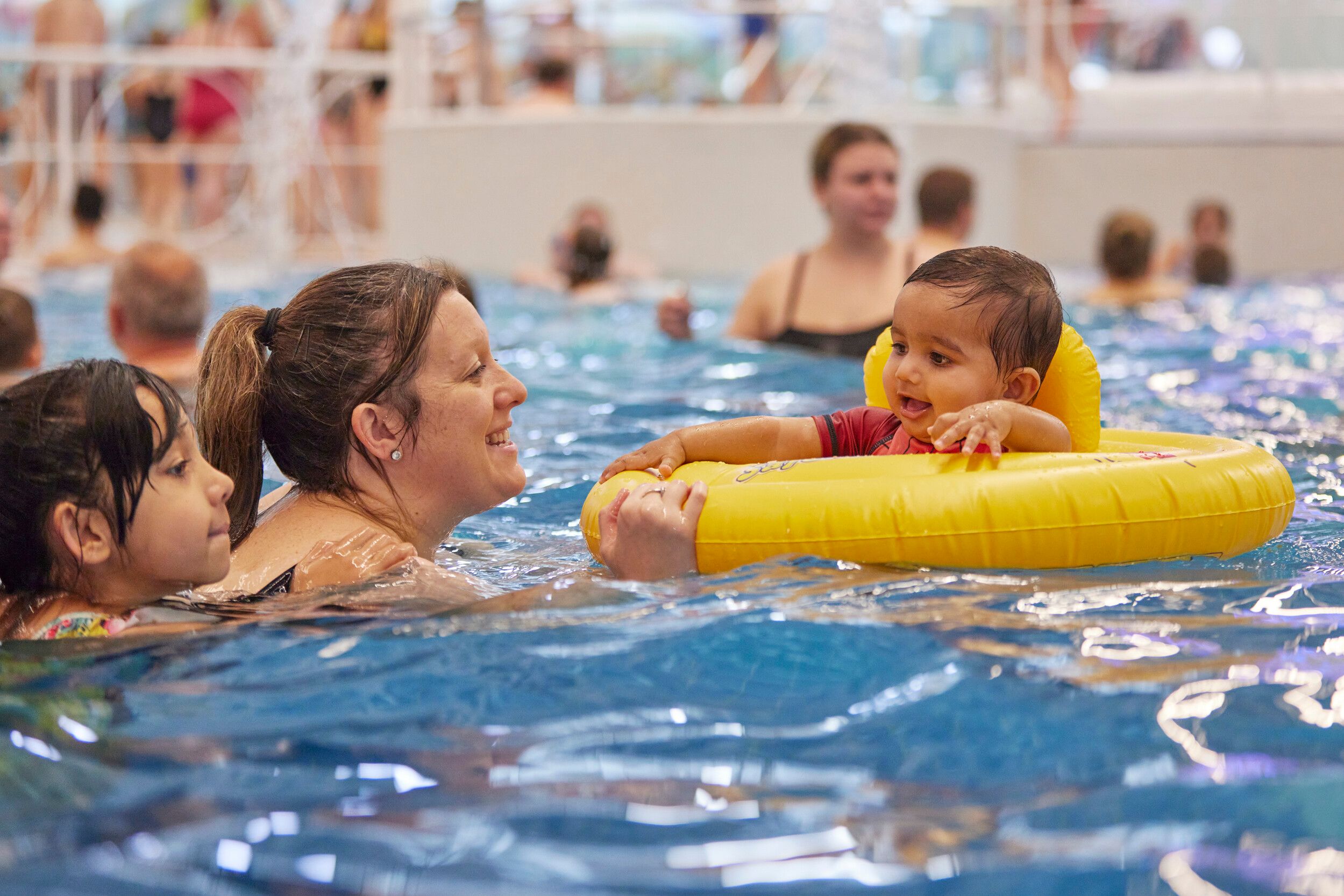 A baby boy riding a yellow float being held by his mother at the pool.