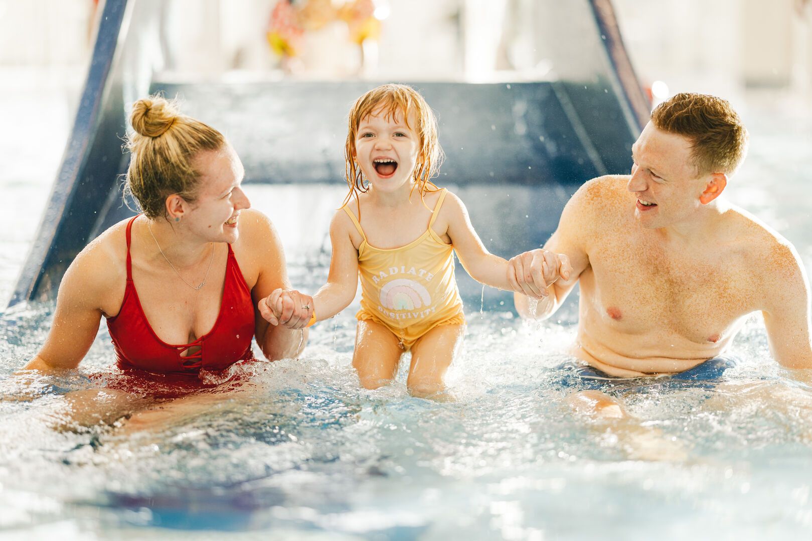 Two parents with their child between them have just gone down a slide together. They are both smiling at their daughter who looks very excited and happy.