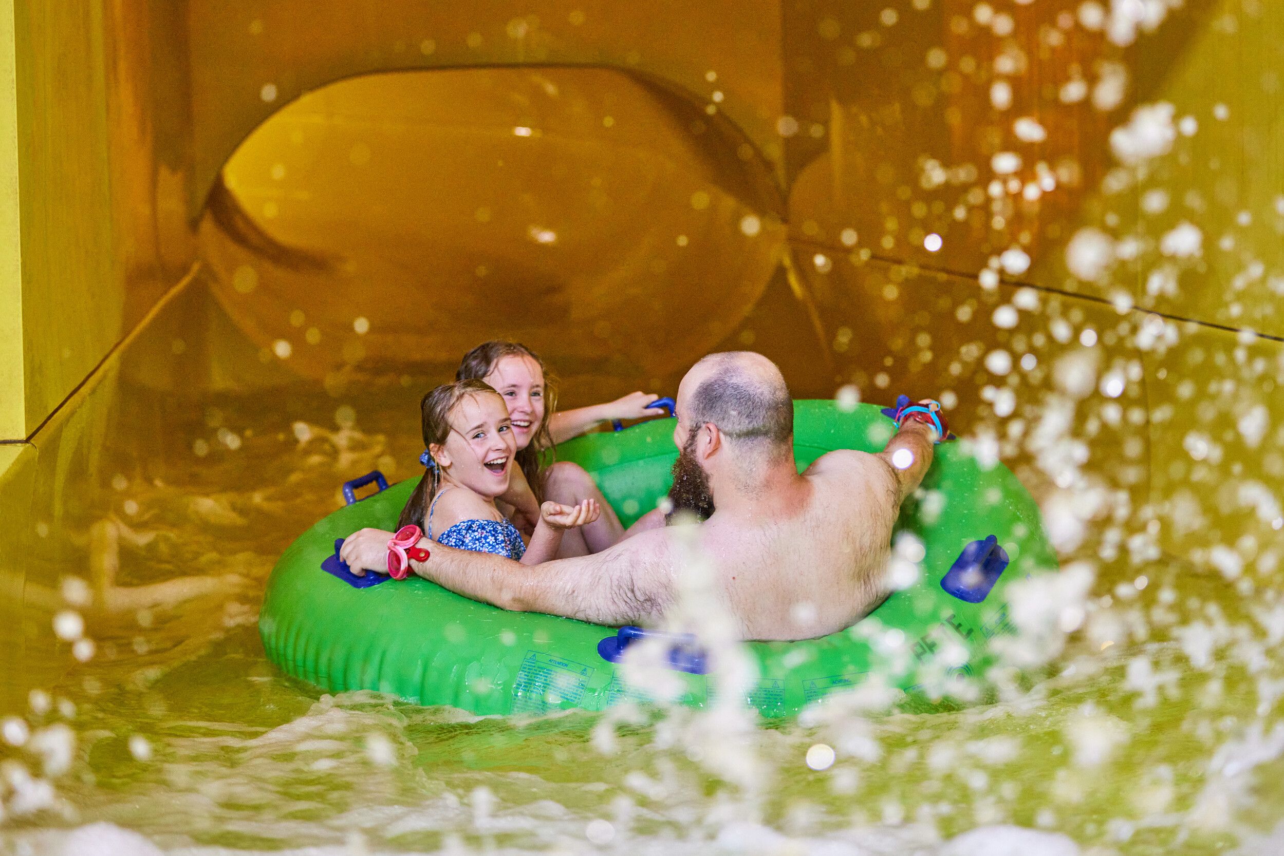 A father and his two daughters riding a green float while going down a yellow water slide.