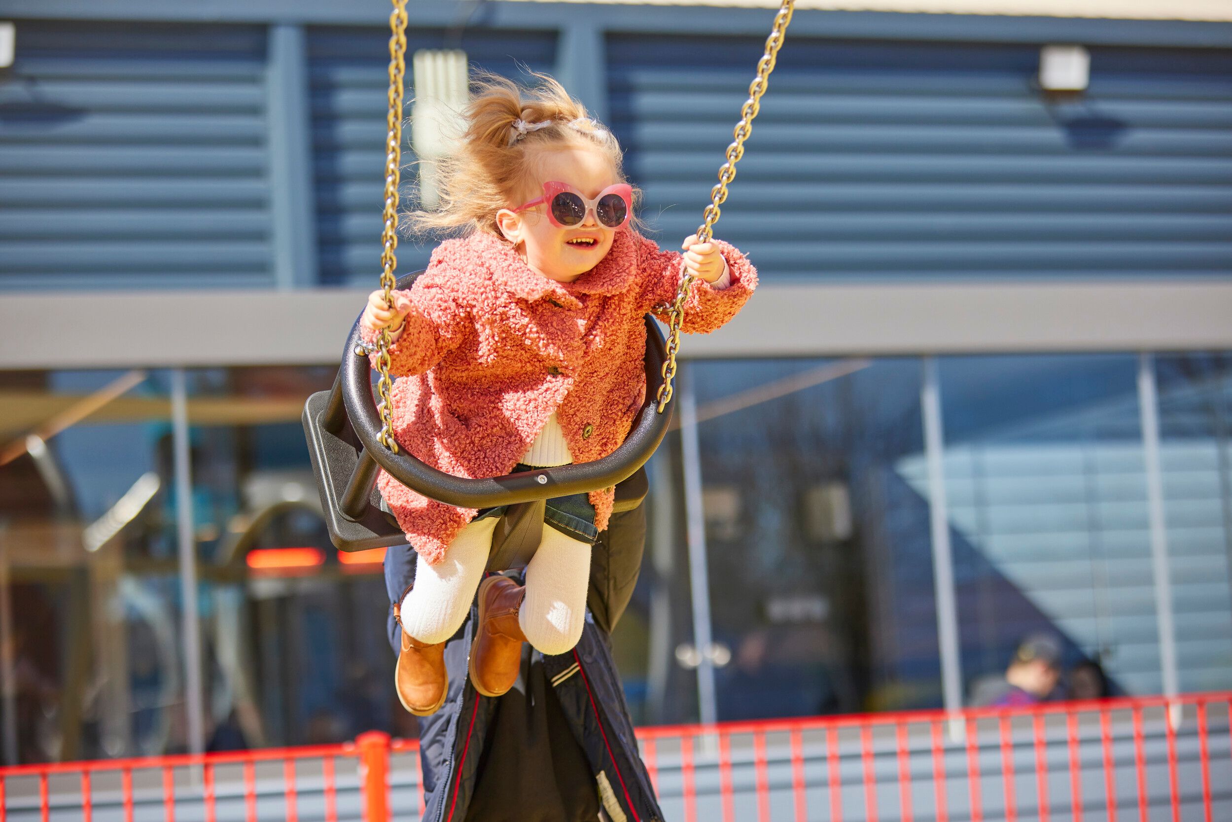 A girl wearing glasses, smiling while riding a swing in Skypark during the day.