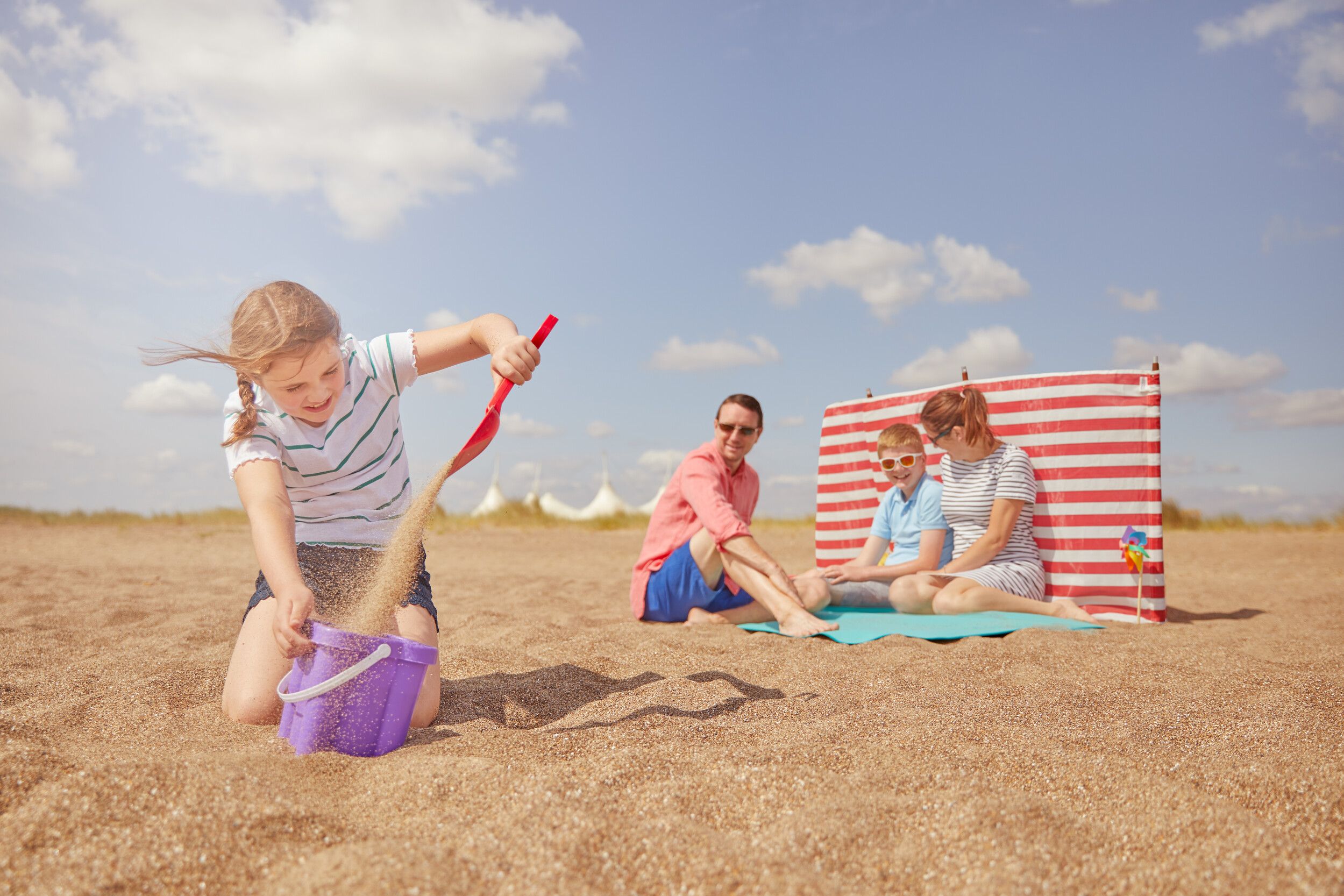 A family sit on the beach together, with a young girl playing with sand in the foreground.