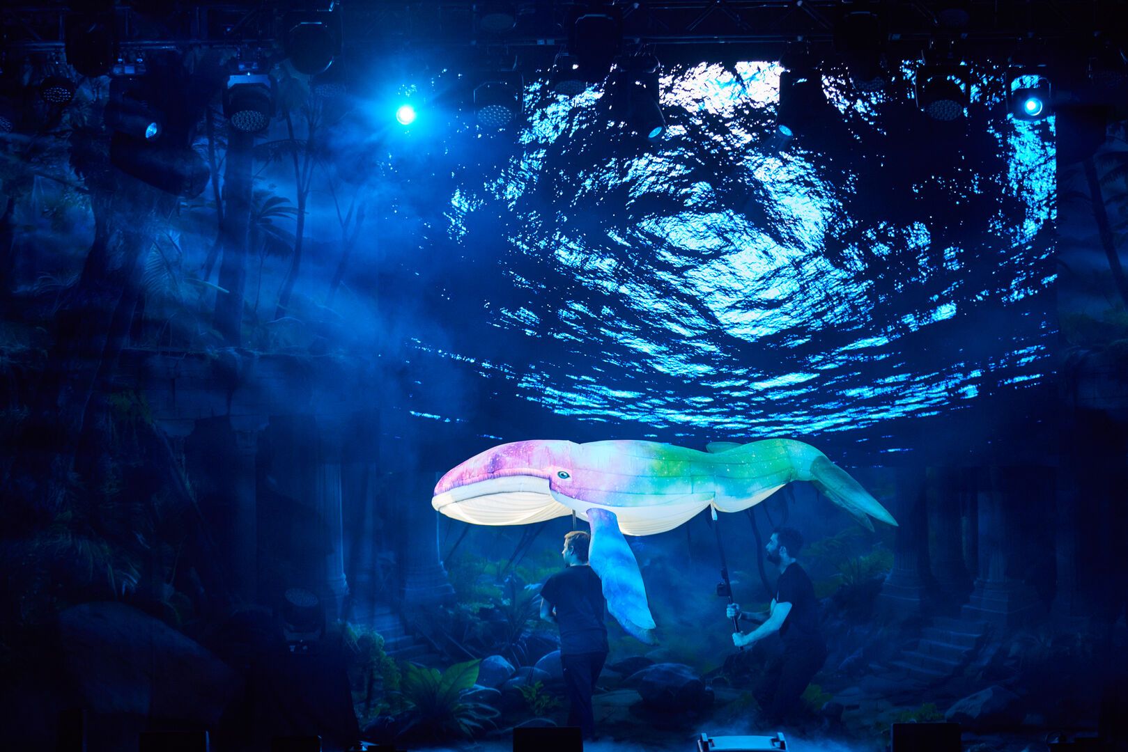 A large man-made whale is shown on stage for the Animals and Mythical Beasts live show at Butlin's. Two stage-hands hold it up, as it is lit in green and blue stage lights.