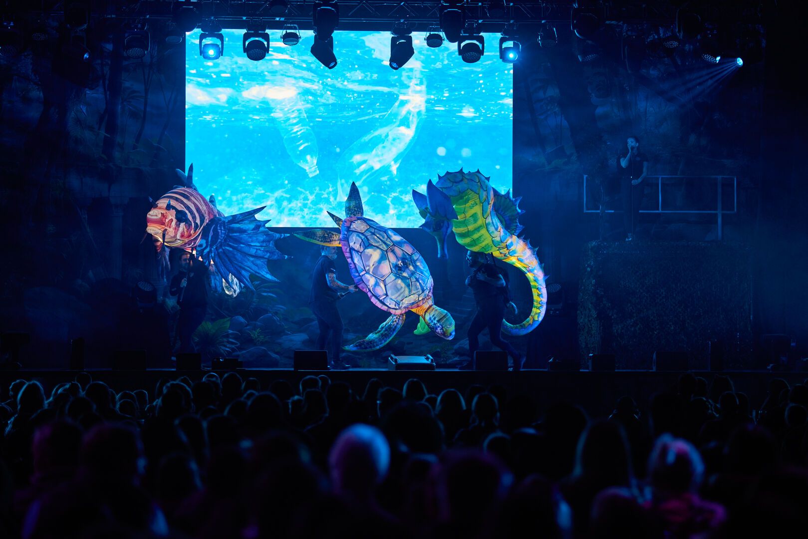 A photo taken at the Animal and Mythical Beasts show. A large crowd can be seen in front of the stage, which is lit in blue lighting. A large seahorse, turtle and Lionfish are being puppeteered on stage.