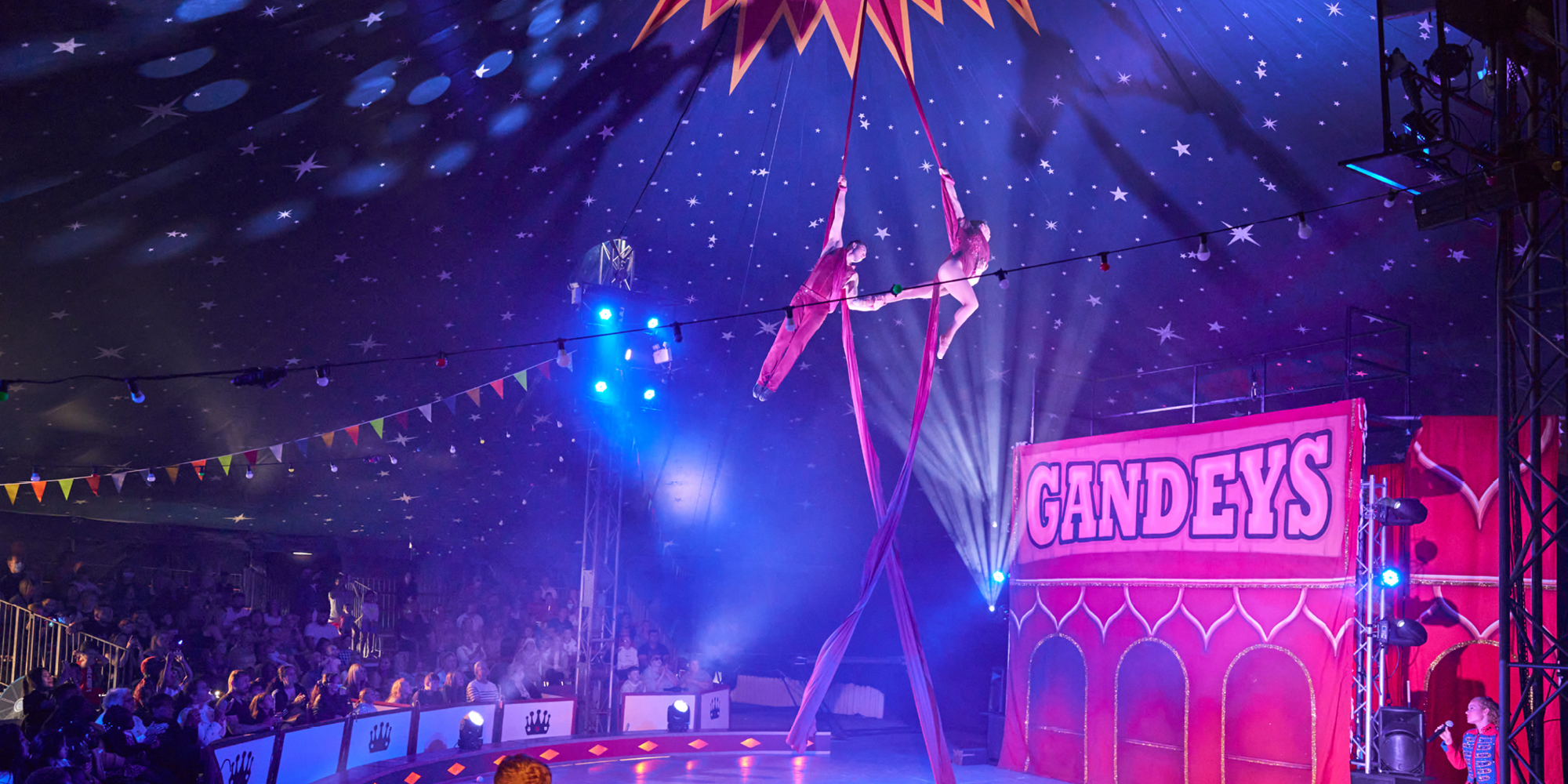 Two performers holding each others hands while performing aerial silk on stage.