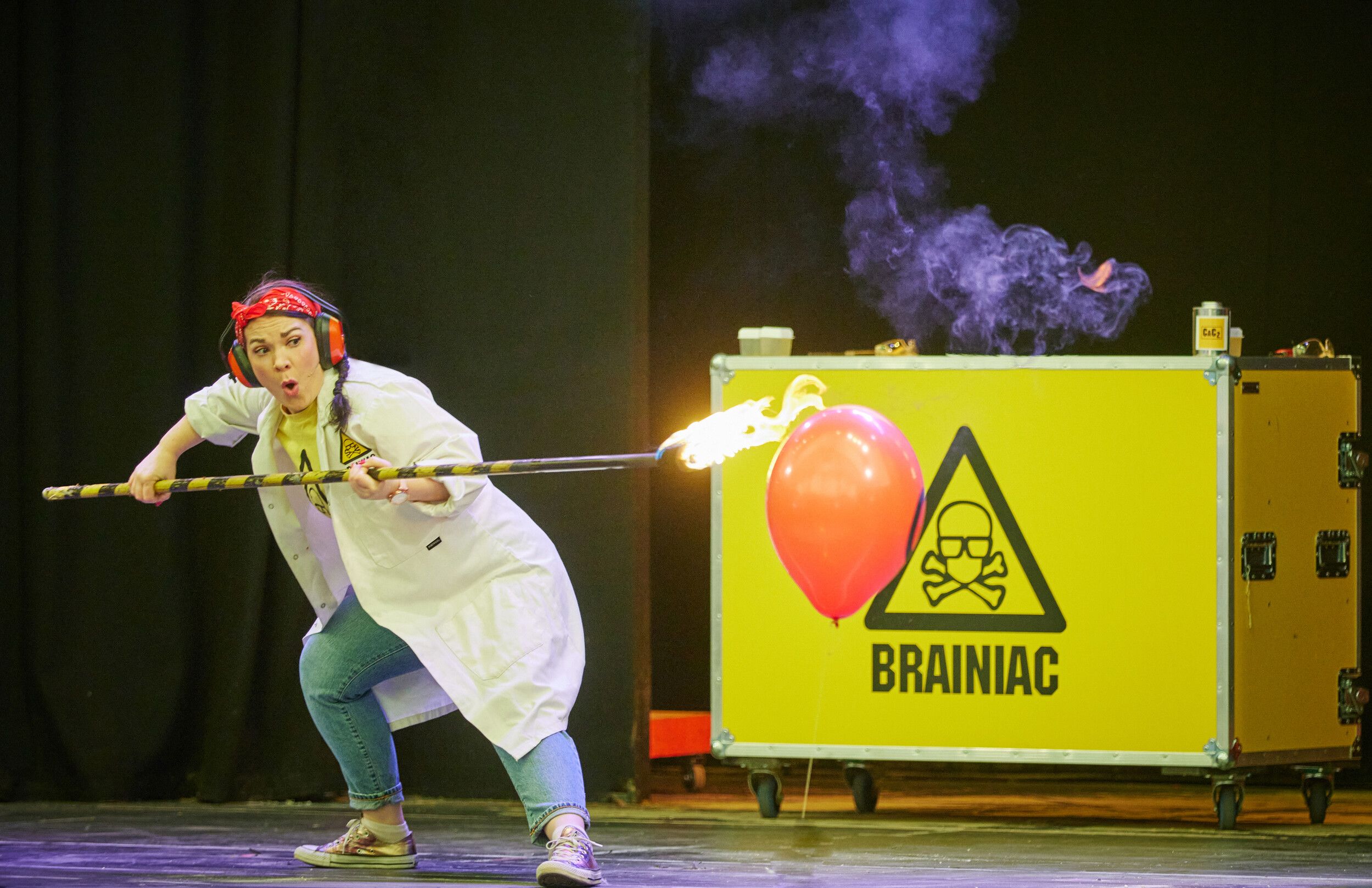 A woman in a white coat experimenting with a fire and a balloon on stage.