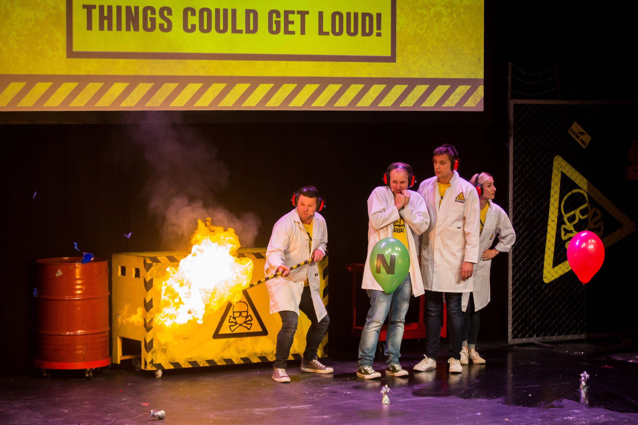 Four people in white coats doing an experiment on stage, the other is holding a stick with a fire.