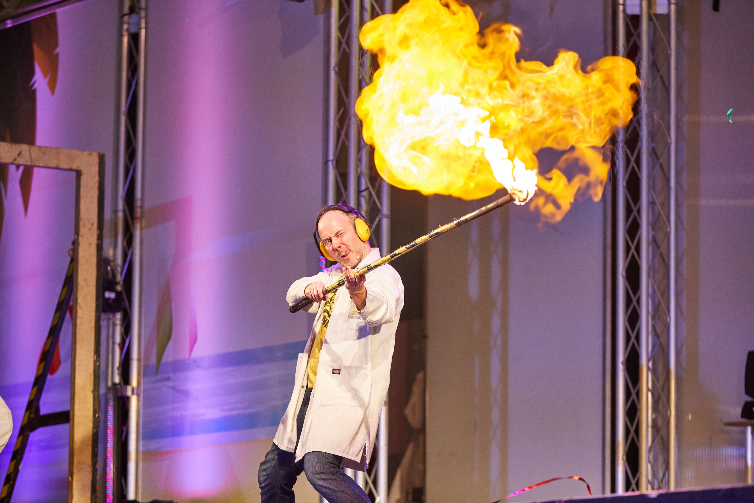 A man in white coat wearing ear muffs while holding a stick with fire on stage.