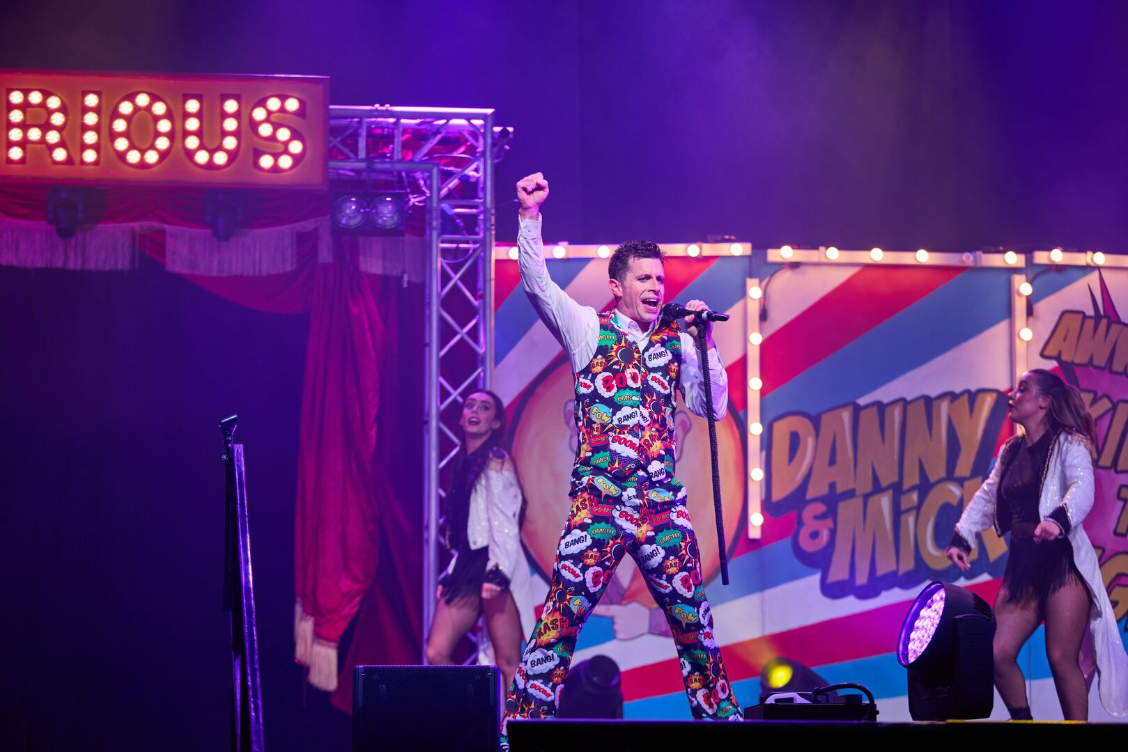A Cirque Du Hilarious performer has one hand in the air, while speaking into a microphone as part of the live show.